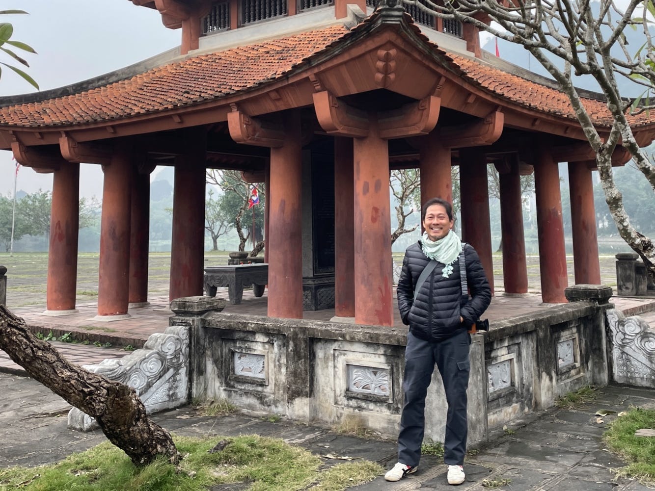 A man wearing a black winter coat posing by a pagoda in Southeast Asia.