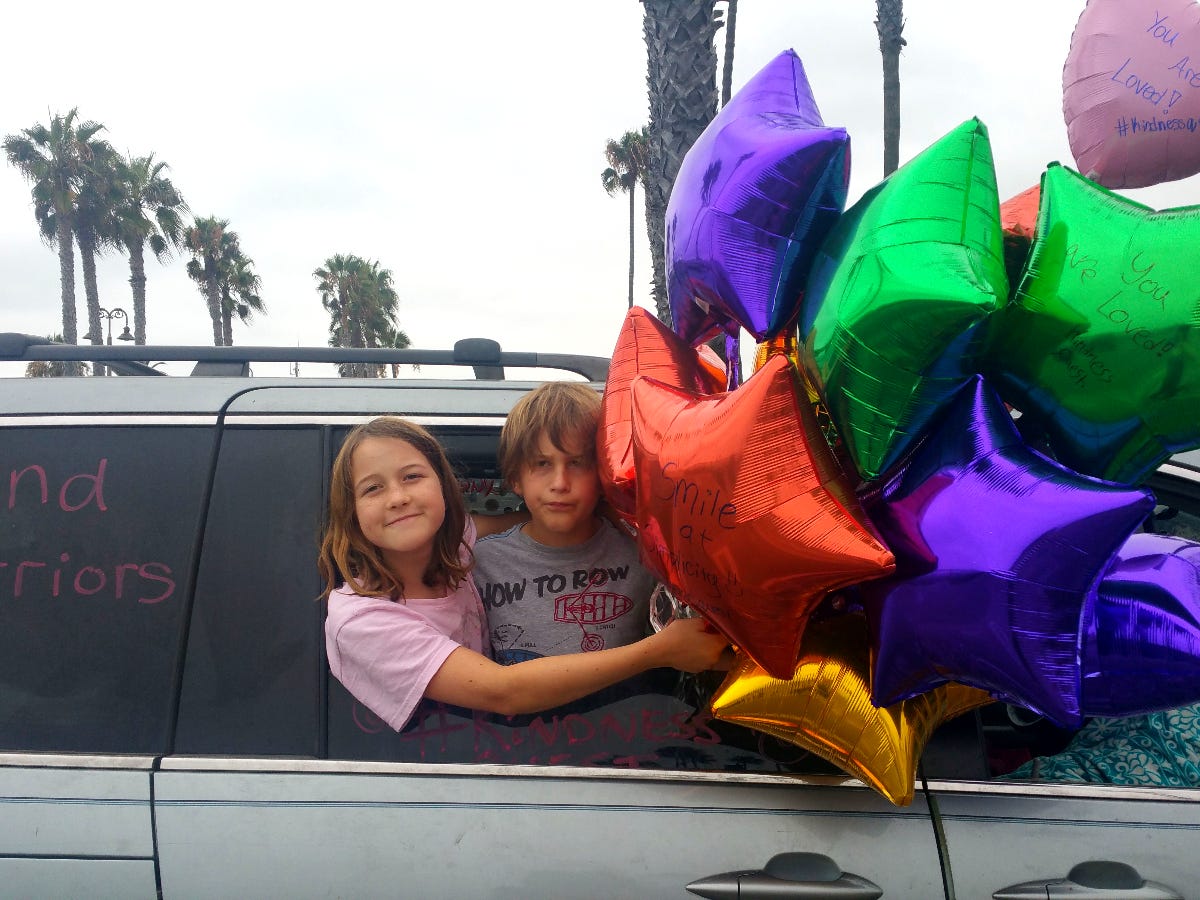 Two children pose while hanging out of the window of a parked minivan. They are holding several star-shaped balloons with kind messages written on. them.