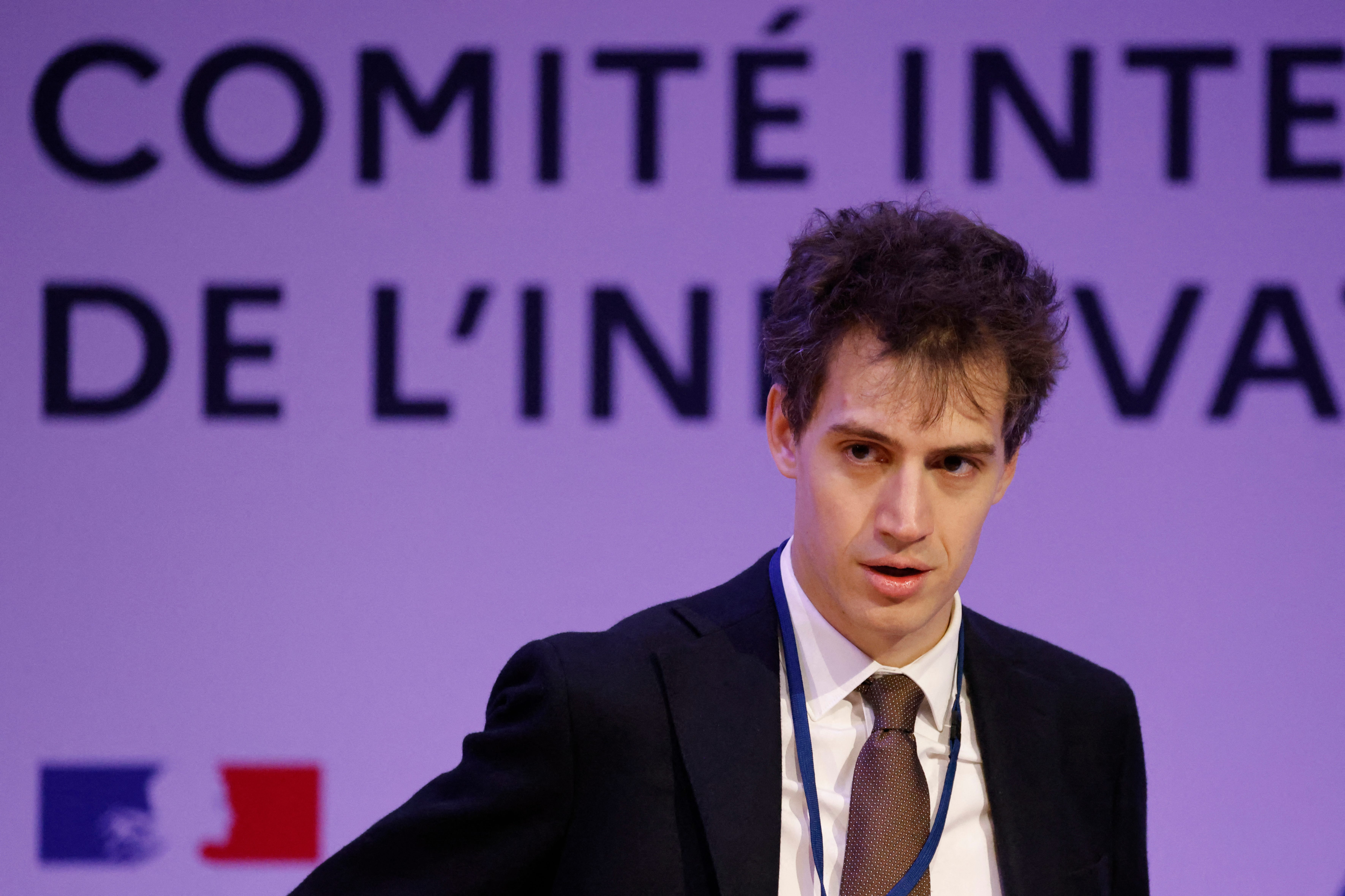 Photo of a man in a suit wearing a lanyard with French text behind him