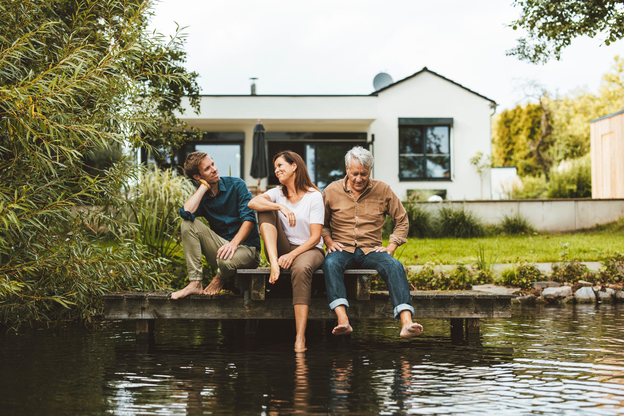 Stock photo of a mother and son talking by senior man sitting on jetty by a lake in the backyard