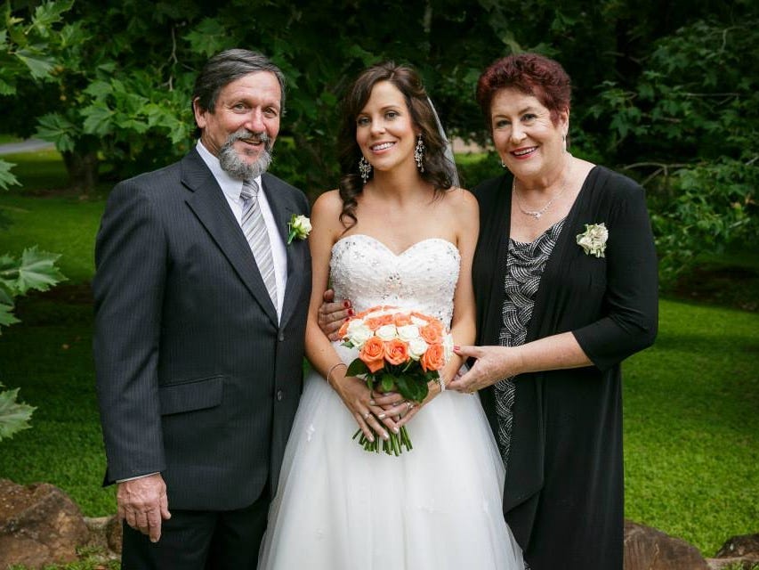 The author is wearing a wedding dress and standing between her parents on her wedding day.