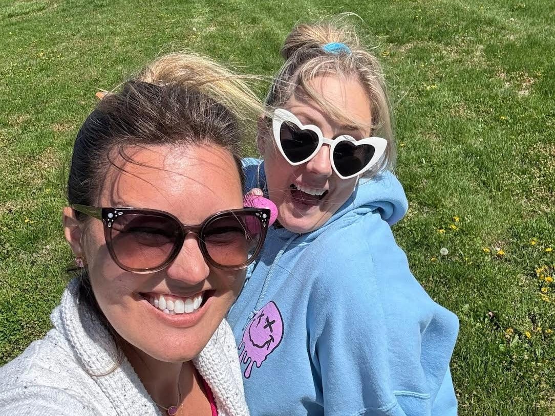 Two women wearing sunglasses pose in a field of grass.