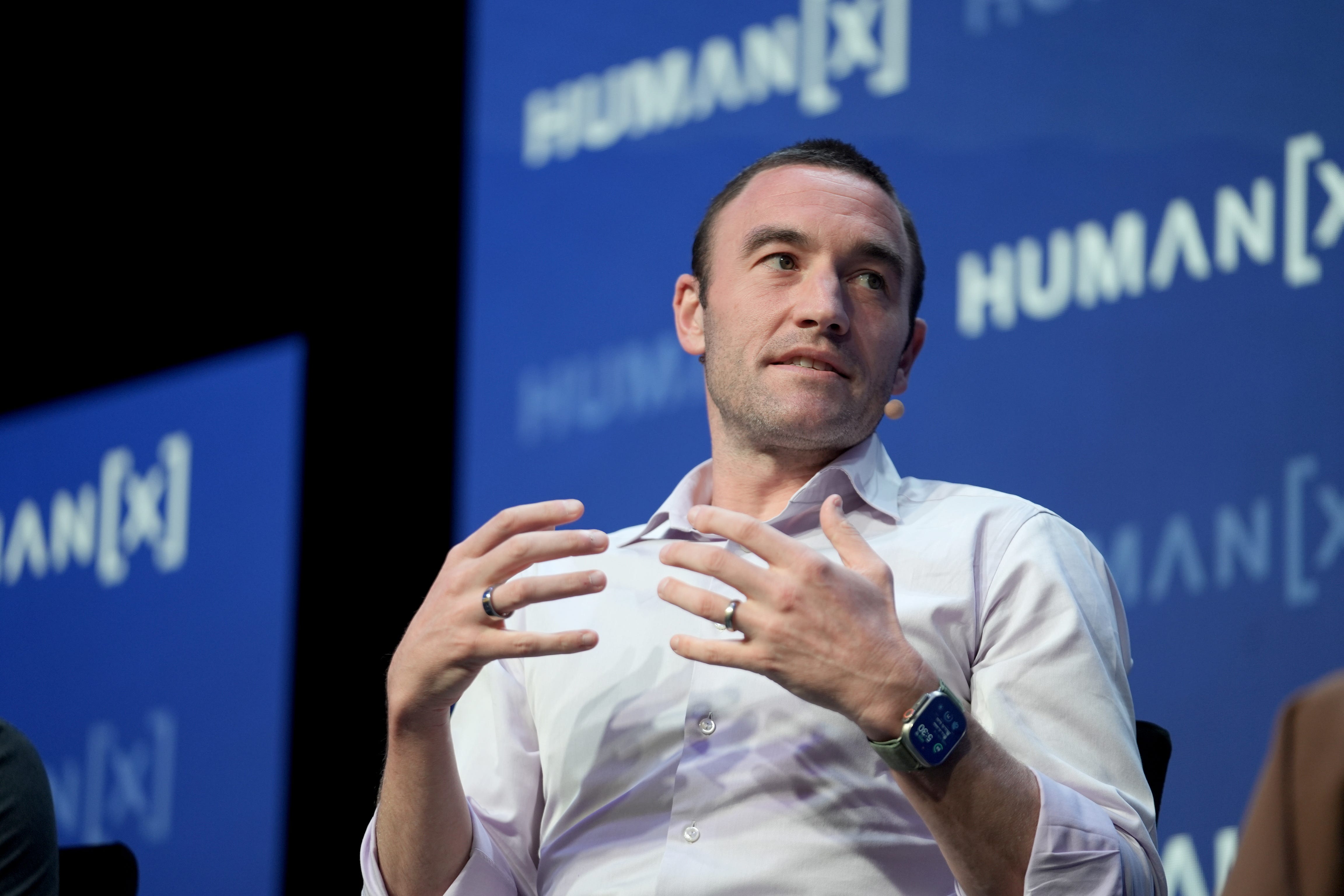 Kevin Weil, CPO of OpenAI, is looking to the left off-camera, and gesturing towards himself with his hands. He is sitting in front of a blue background and wearing a white button-down shirt. He is mid-speech.