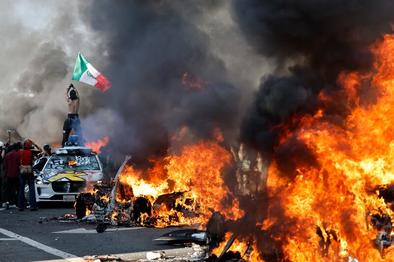 A protestor holds up a Mexican flag as burning cars line the street on June 08, 2025 in Los Angeles, California. Tensions in the city remain high after the Trump administration called in the National Guard against the wishes of city leaders following two days of clashes with police during a series of immigration raids.