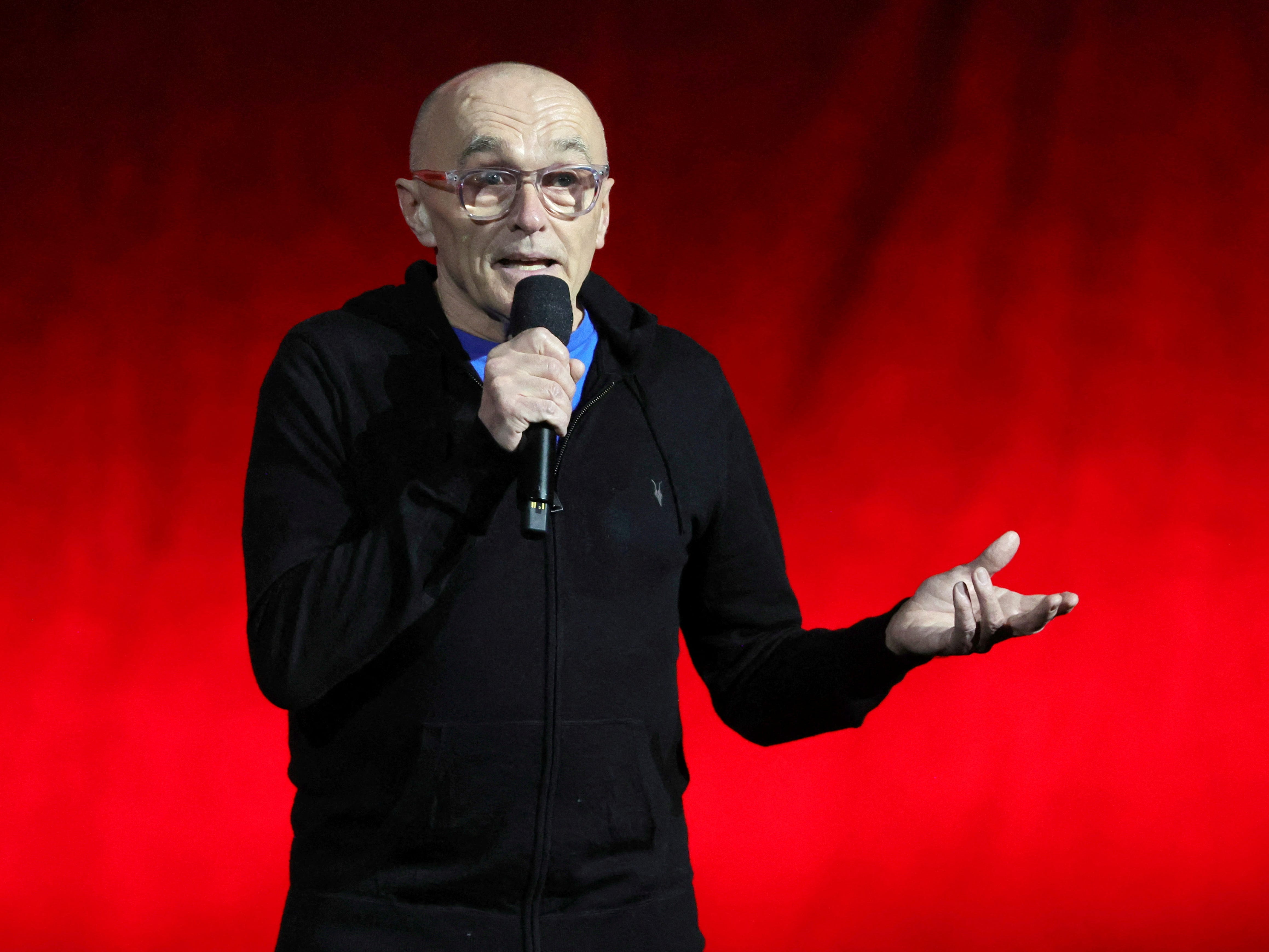 A bald man in a black hoodie holding a black microphone. HE's wearing a blue t-shirt and clear glasses. He's stood against a bright red backdrop.