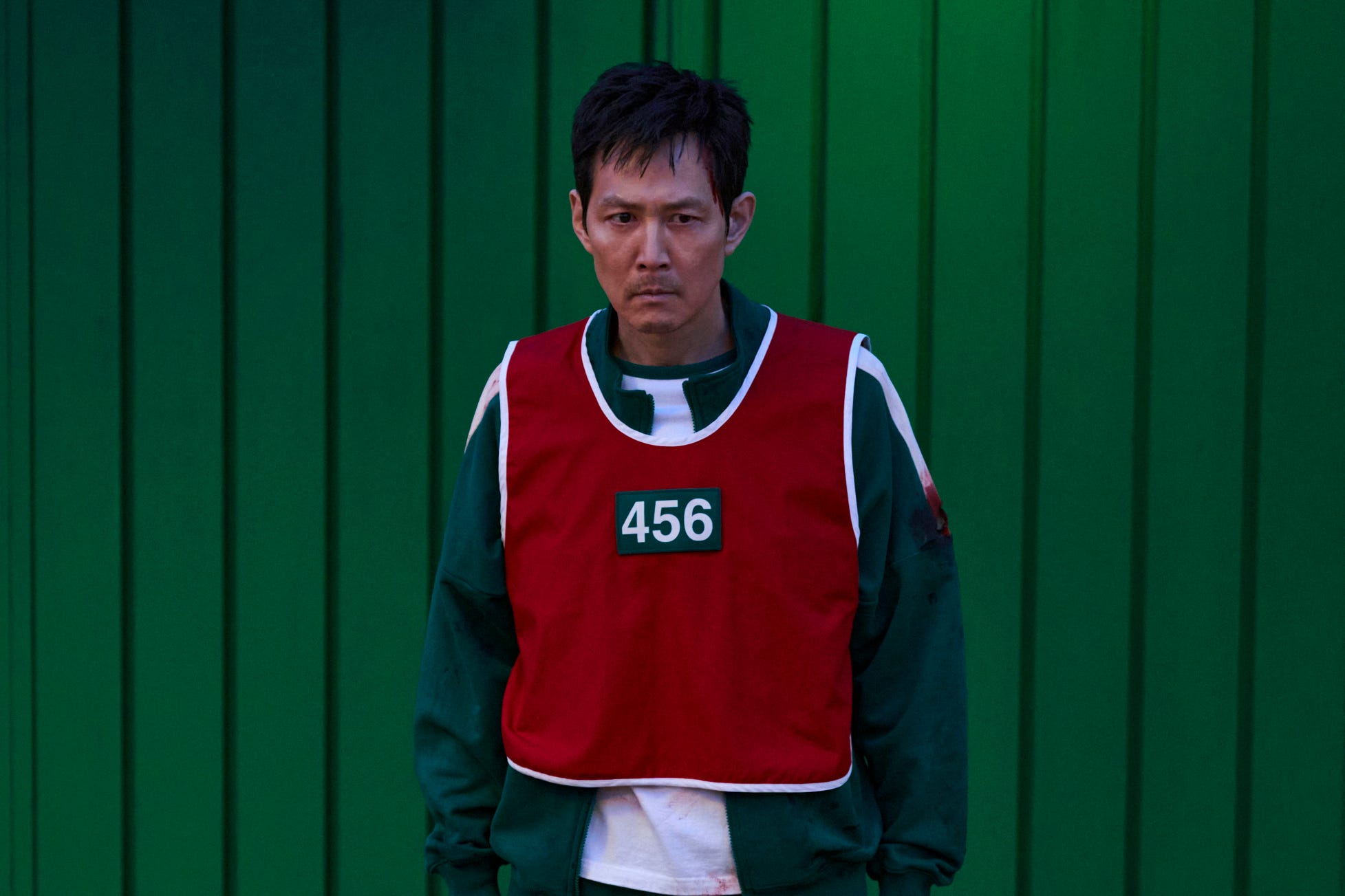 A still of "Squid Game" showing Lee Jung-jae in a bloody green tracksuit and red vest.