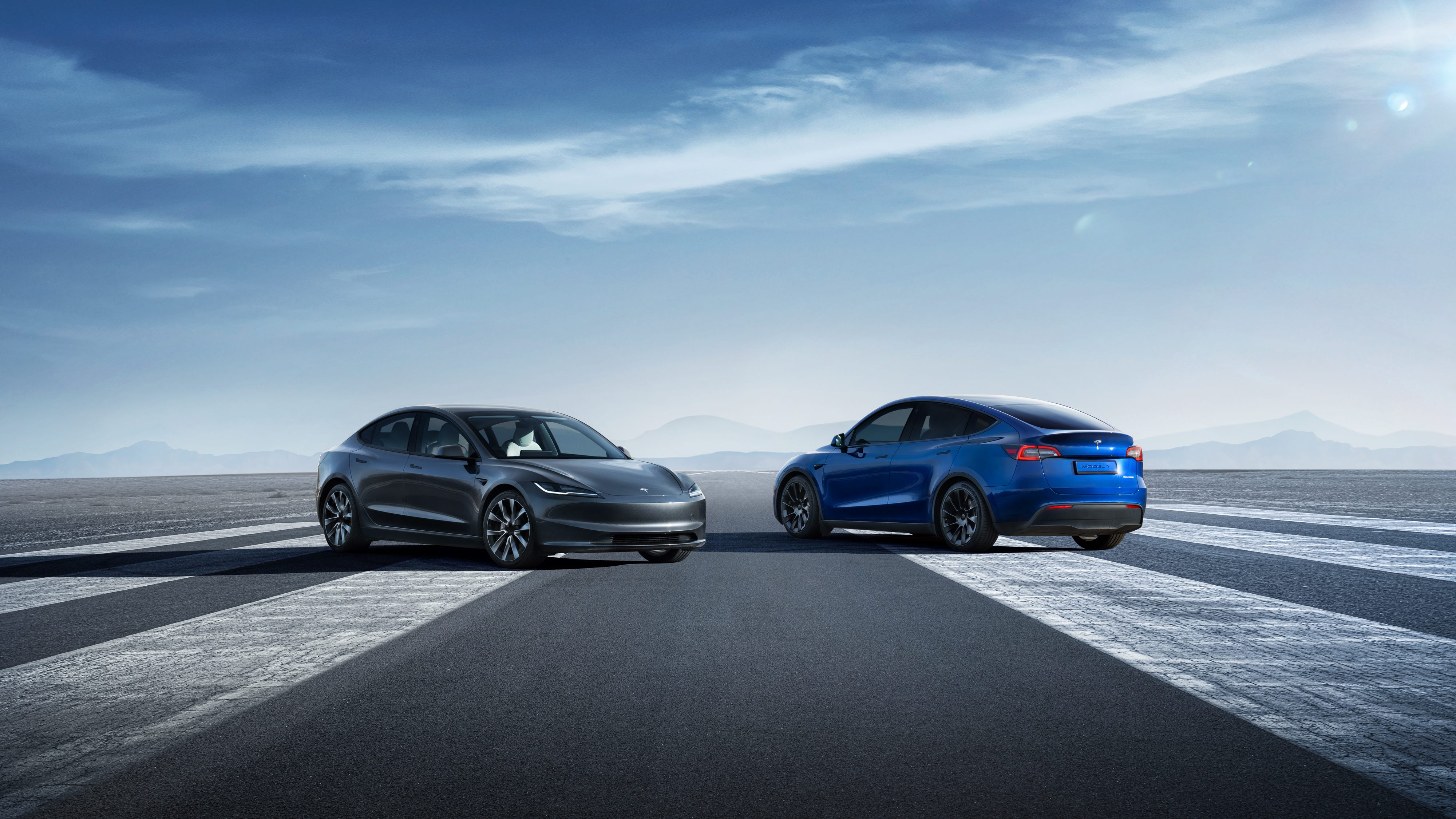 The right front of a gray Tesla Model 3 Sedan and the left rear of a blue Tesla Model Y SUV parked next to one another on a runway.