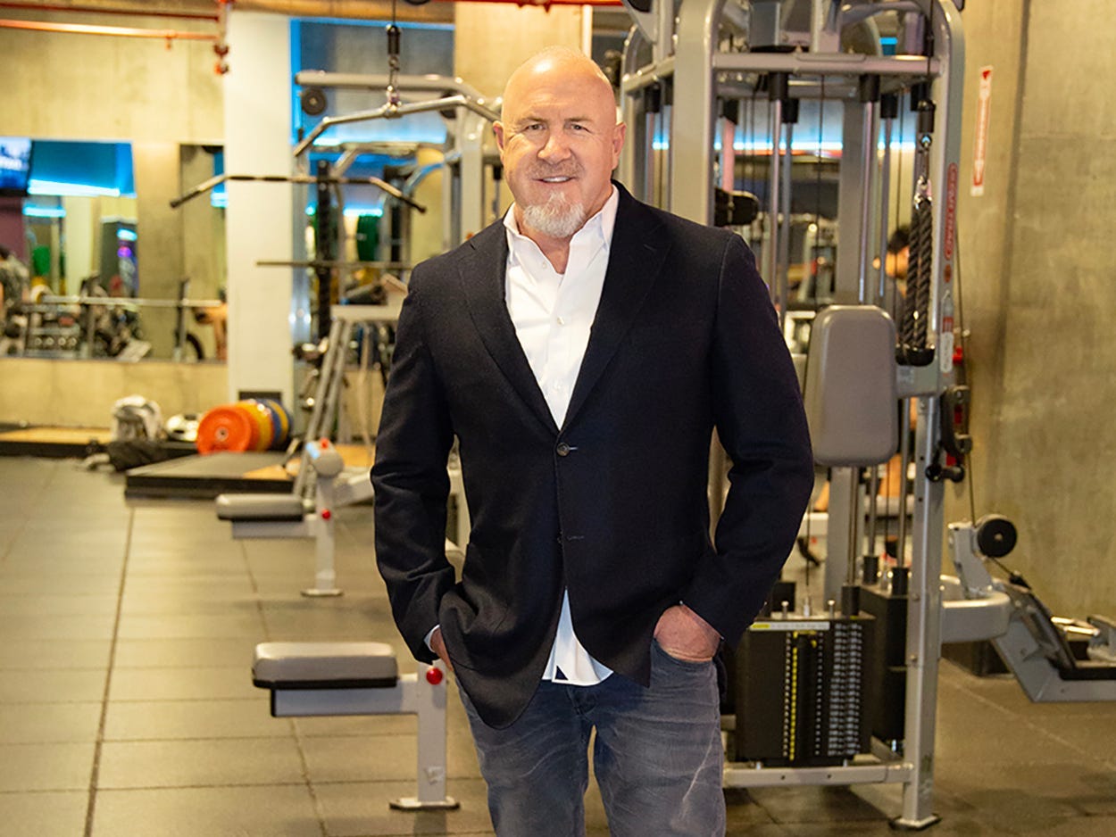 a man in a formal shirt and blazer and jeans smiling at the camera in a gym