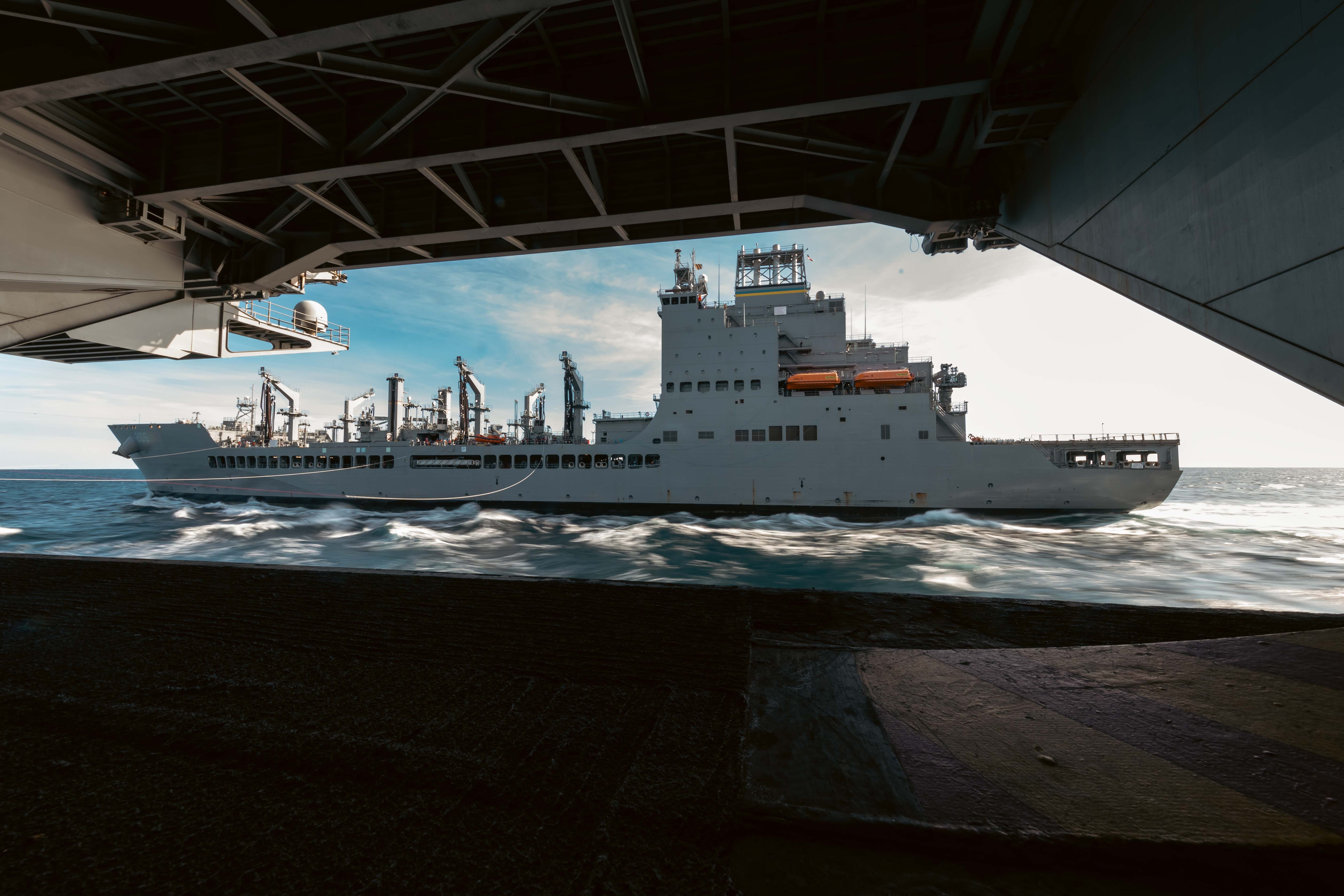 A view of the USS Harvey Milk John Lewis-class replenishment oiler is seen from inside a ship on blue ocean water with a cloudy blue sky in the background.