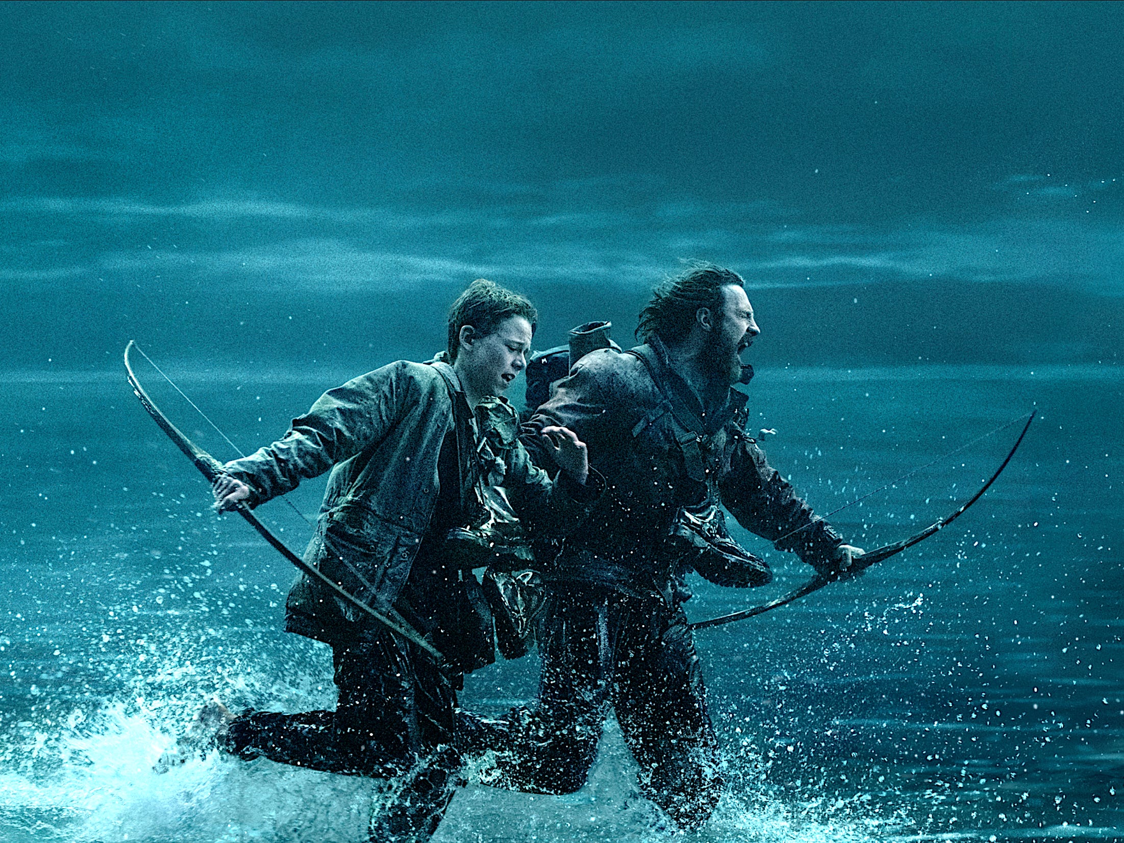 A teenage boy and a man run through the sea at night. They're both dressed in dark clothing and have their boots tied around their necks. They're both holding wooden bows.