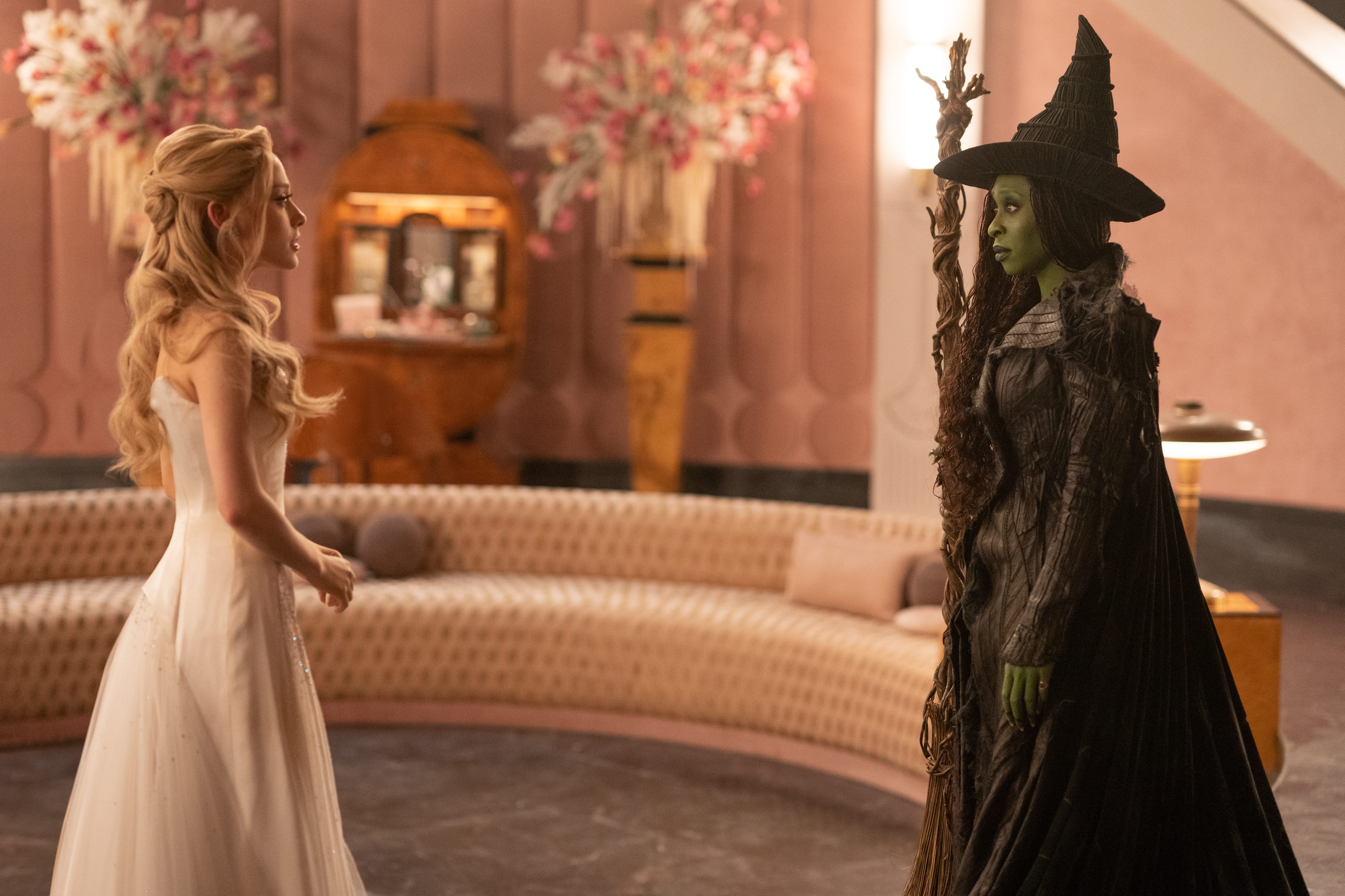 A still for "Wicked: For Good" showing Ariana Grande in cream dress looking at Cynthia Erivo in green makeup and black witch clothing.