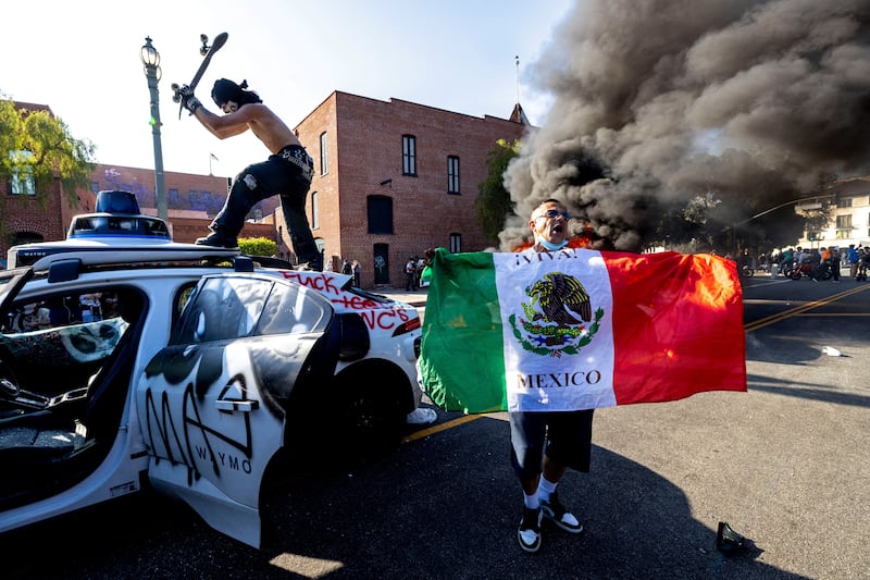 A demonstrator hits a Waymo self-driving car with his skateboard as another burns at rear, during a protest against federal immigration sweeps, in downtown Los Angeles.