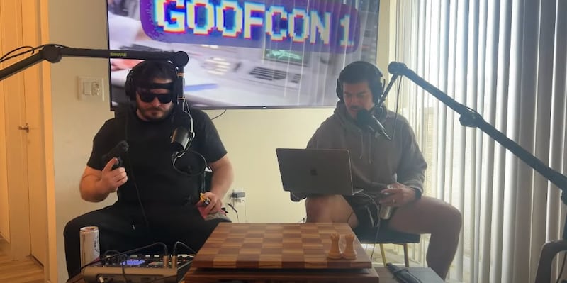 Two men sitting in front of microphones in a living room with bad stage lighting.