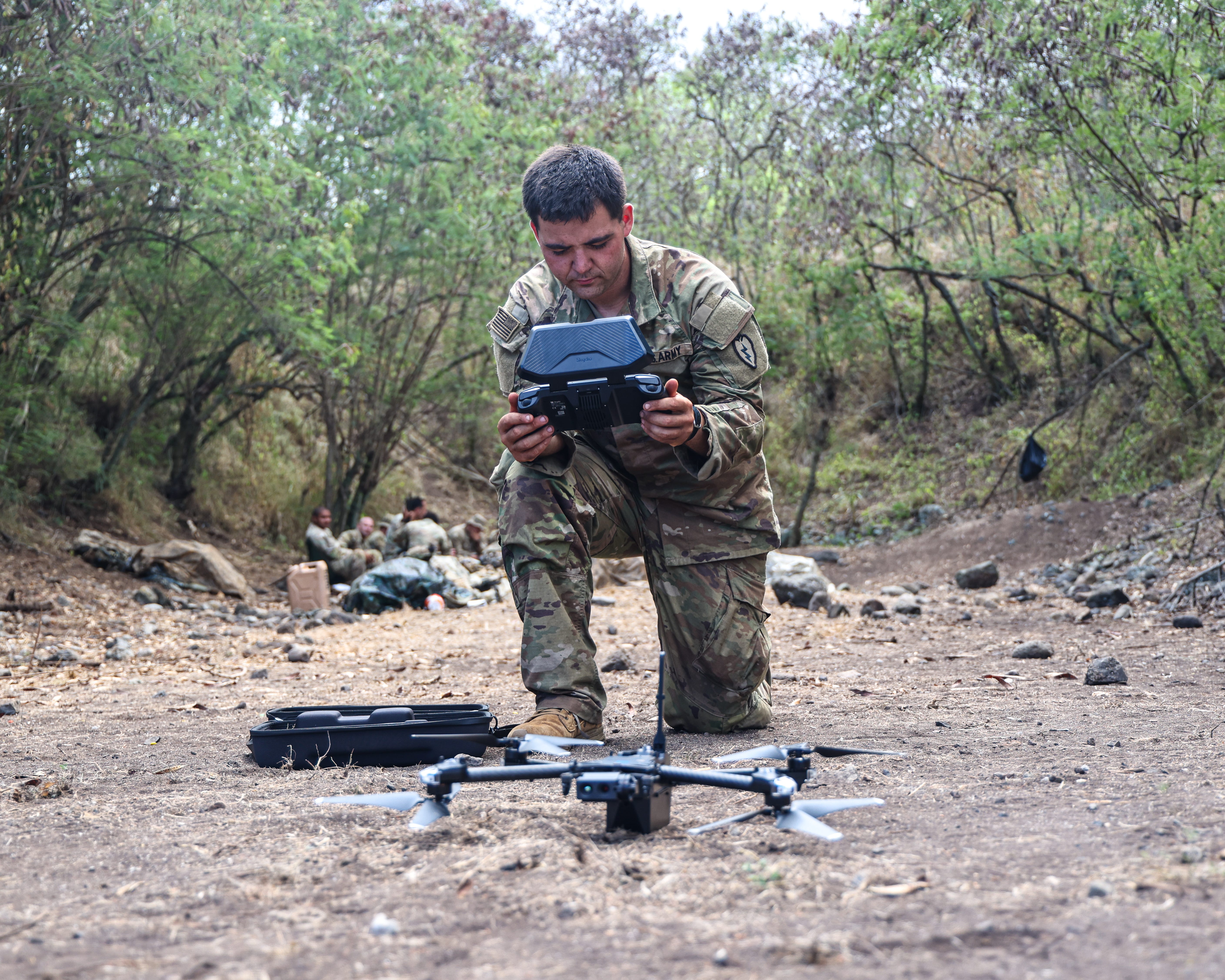 A man wearing an Army camouflage uniform kneels on one knee holding a drone controller in his hands. A quadcopter drone is on the ground in front of him. Behind the man is a line of green trees.
