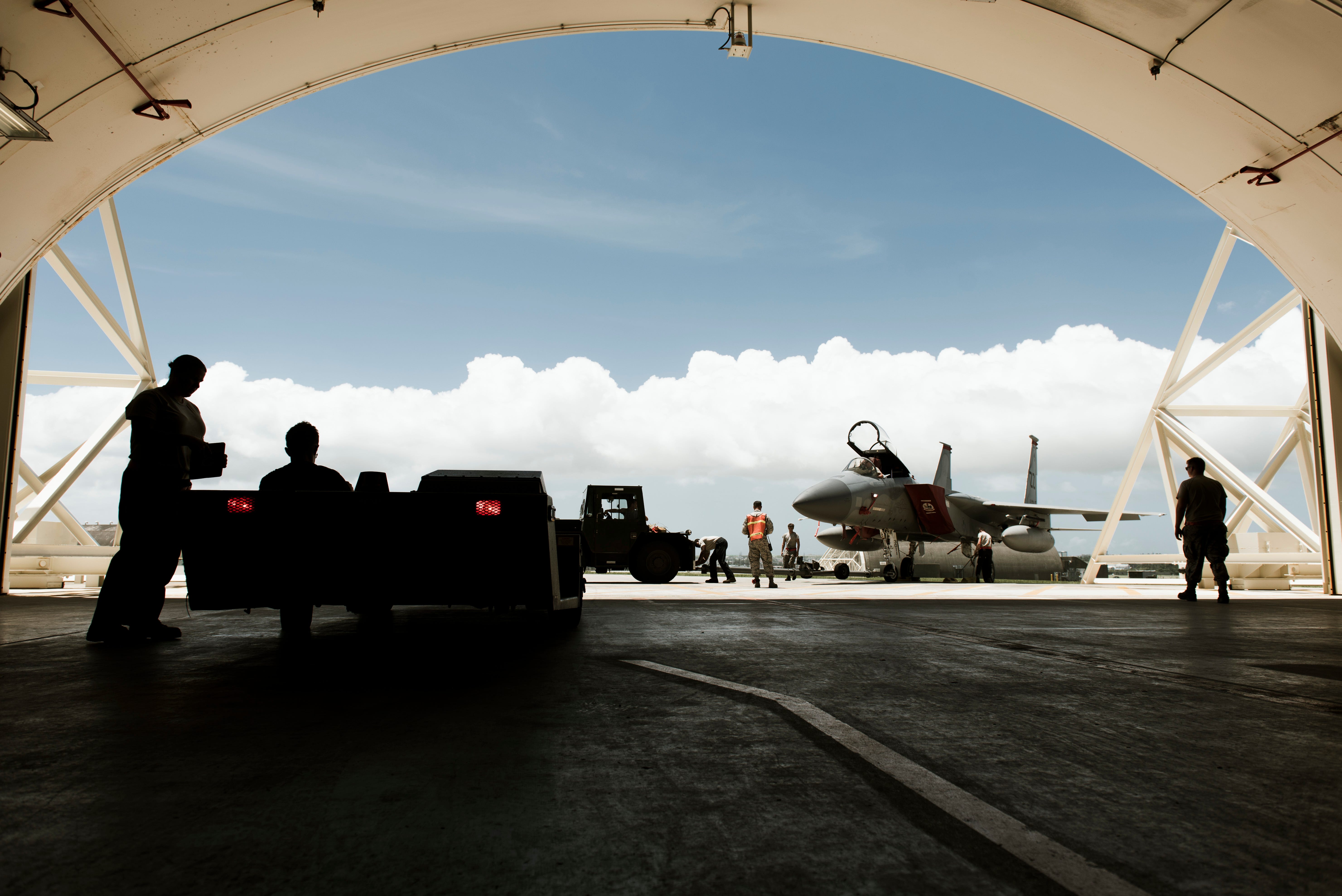Inside an airbase shelter, an aircraft sits on a runway and people stand around it. The background is a blue sky with a line of clouds.