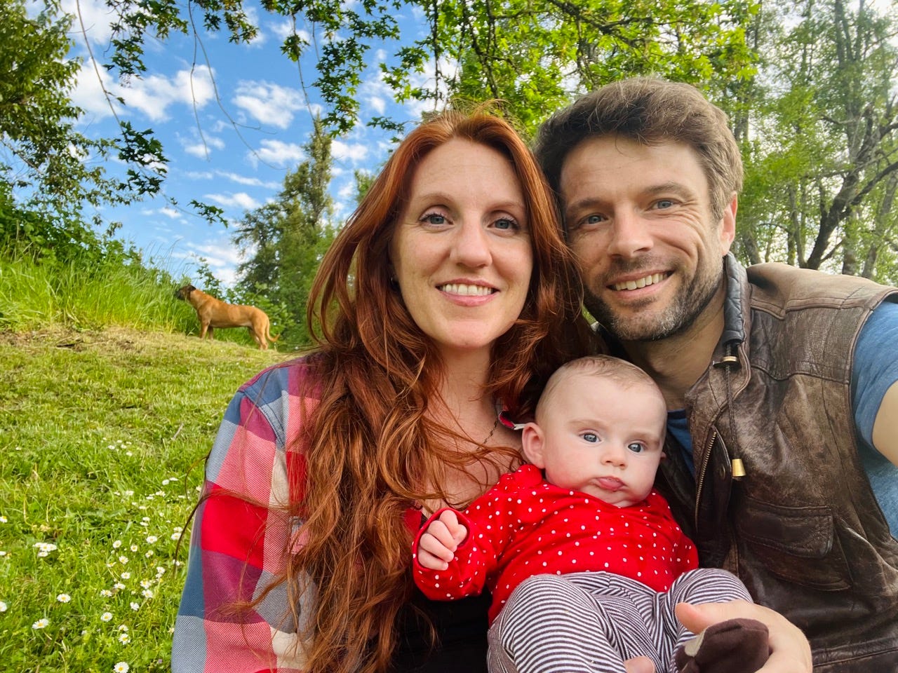 Author Lizabeth Yandel, her partner, and their daughter in front of a grassy field with a dog in the distance