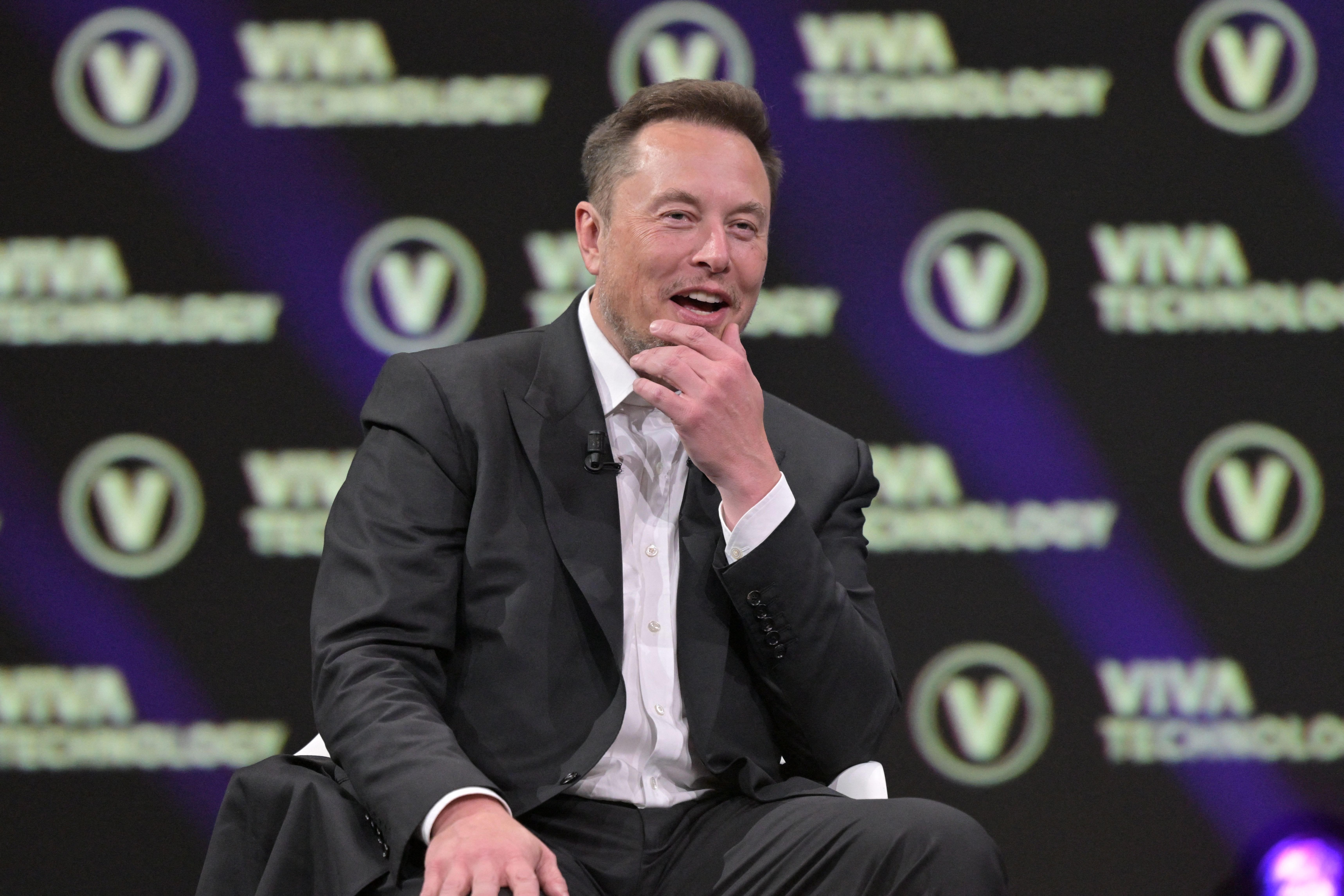Elon Musk in a gray jacket and white dress shirt reacts in front of a purple and yellow step-repeat background.