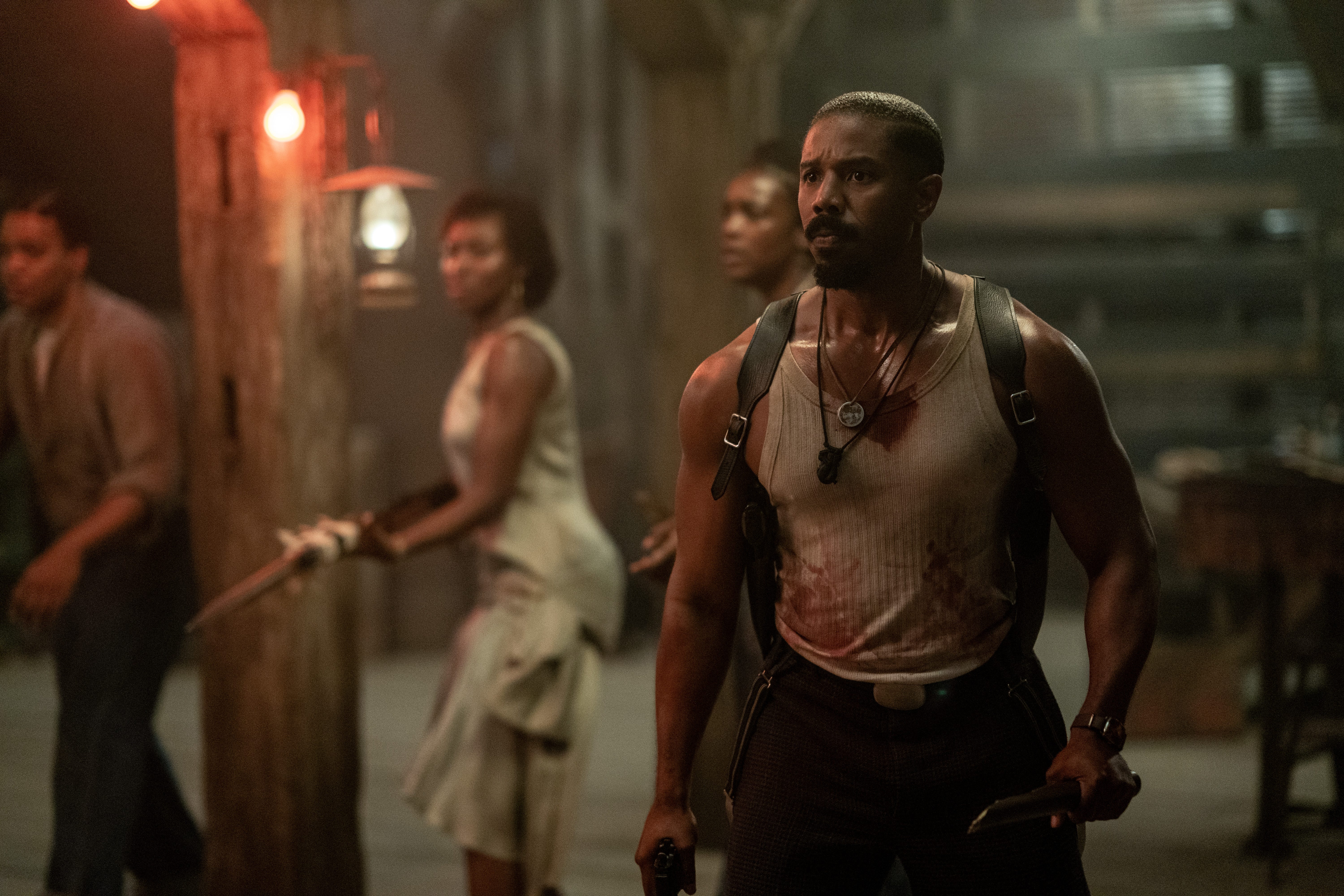 A still of "Sinners" showing Michael B. Jordan in a bloody vest, holding a broken wooden stick and gun with other people with weapons in the background.