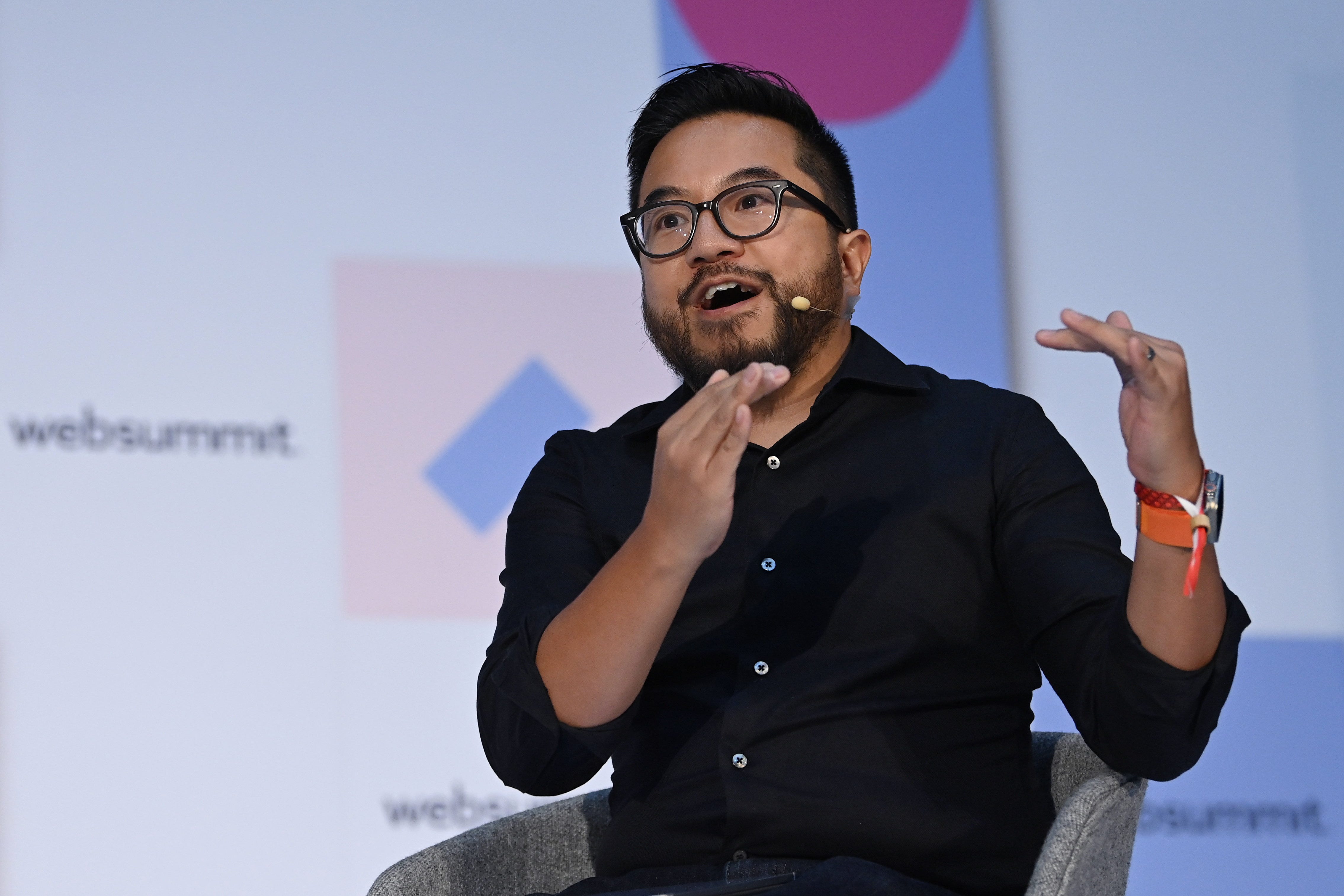 Garry Tan, the CEO of Y Combinator, is gesticulating with his hands and is mid-speech. He is wearing a smart watch with an orange band, a black collared button up, and blue jeans. He is sitting in a grey chair in front of a background with text that reads, "websummit."