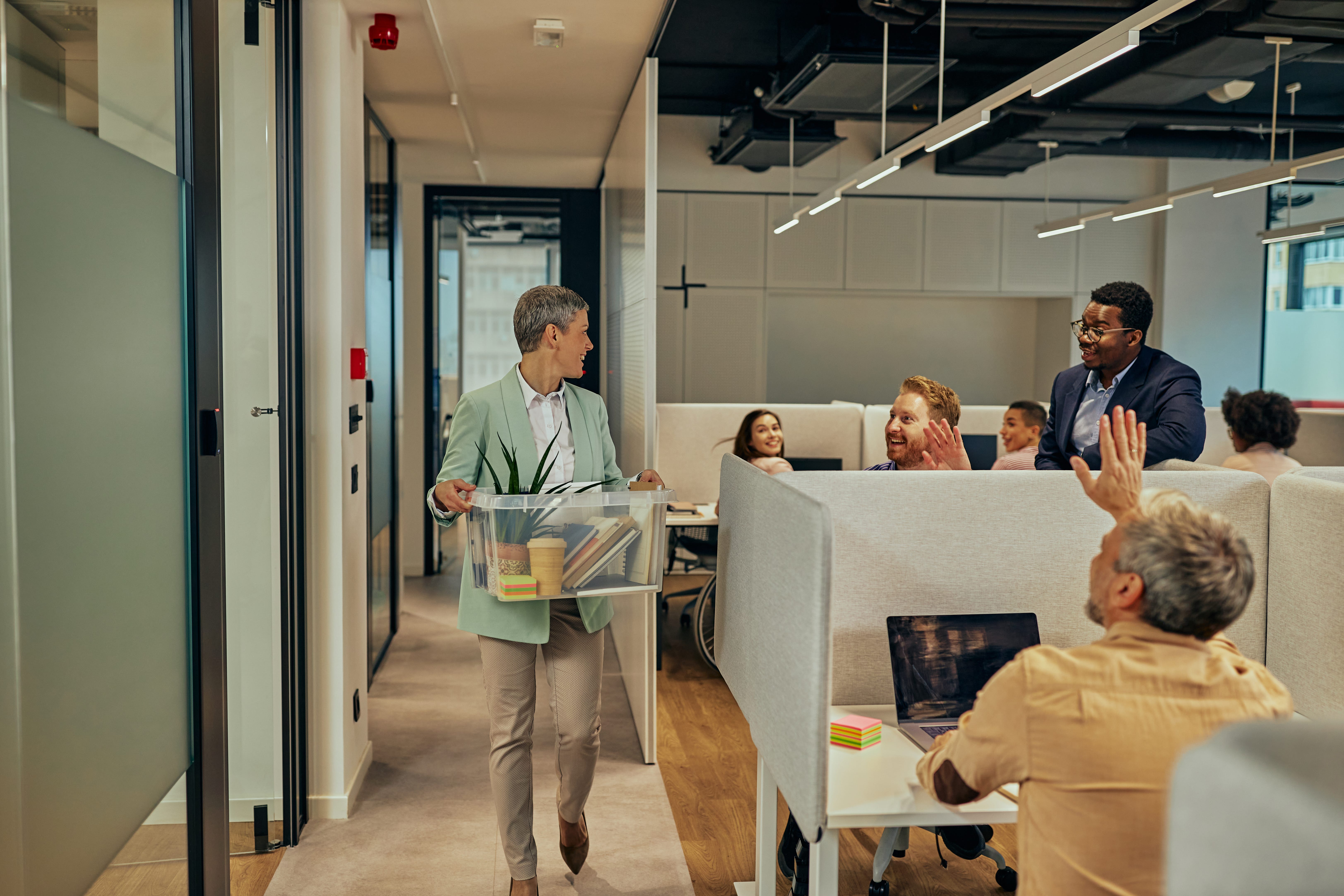 A woman greets employees carrying a box of things in an office.