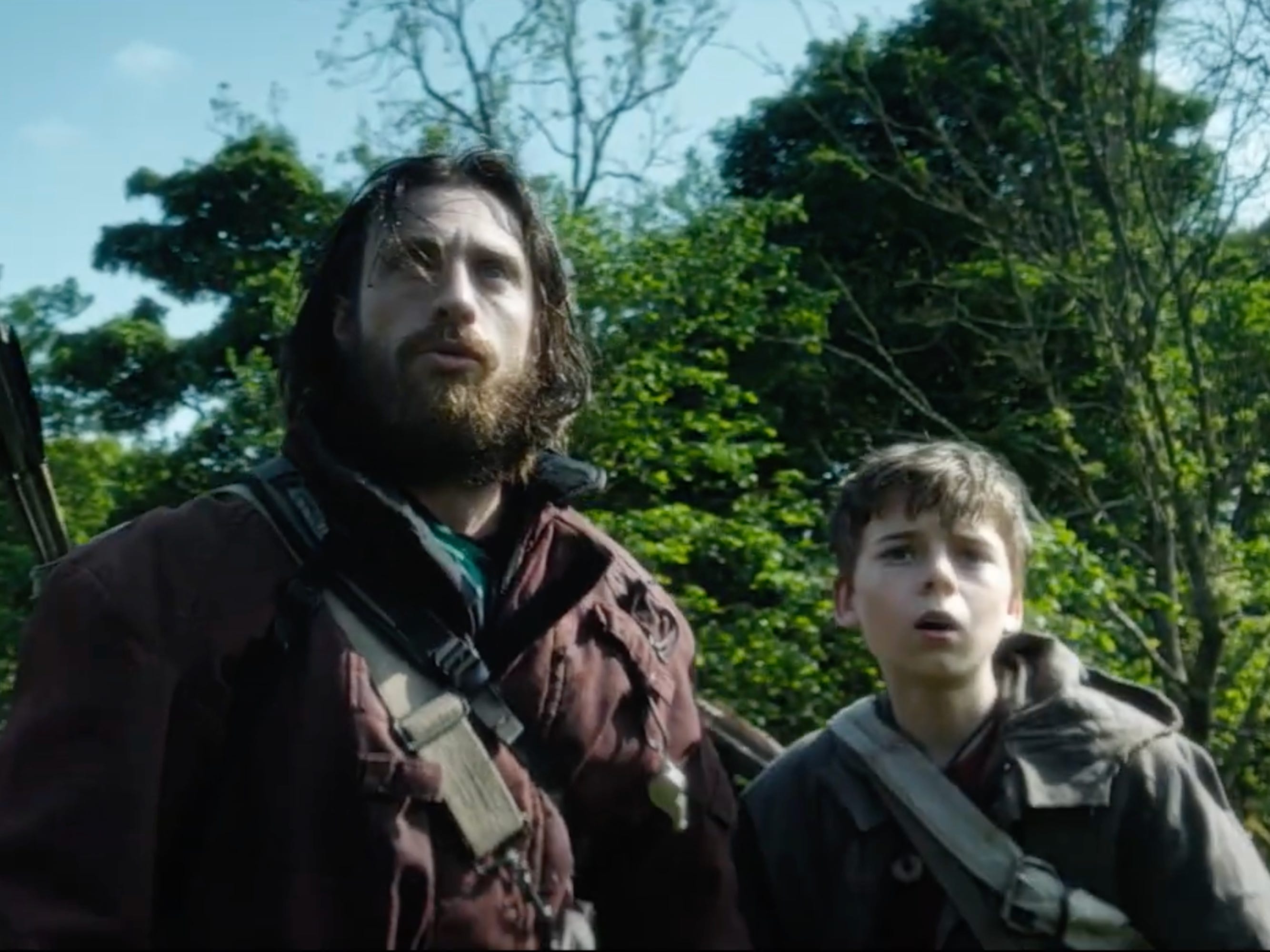 A man and a boy standing in a woodland area. On the left, the man has long brown hair and a long brown beard. He's wearing a burgundy coat with a quiver of arrows strapped to his back. On the right, the young boy wears a black hodded coat and has a white bag strap across his chest.