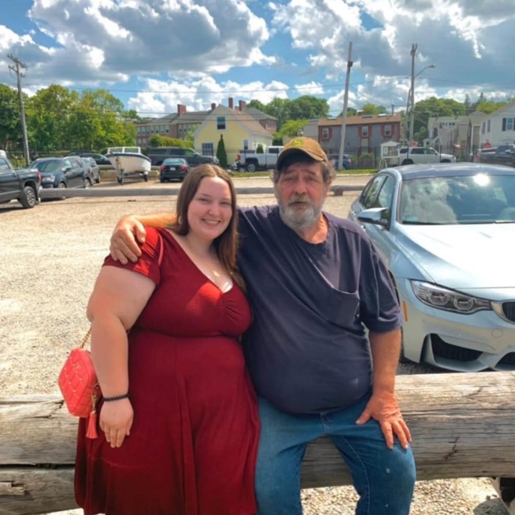 A young woman in a red dress and an older man with a beard sitting on a log.