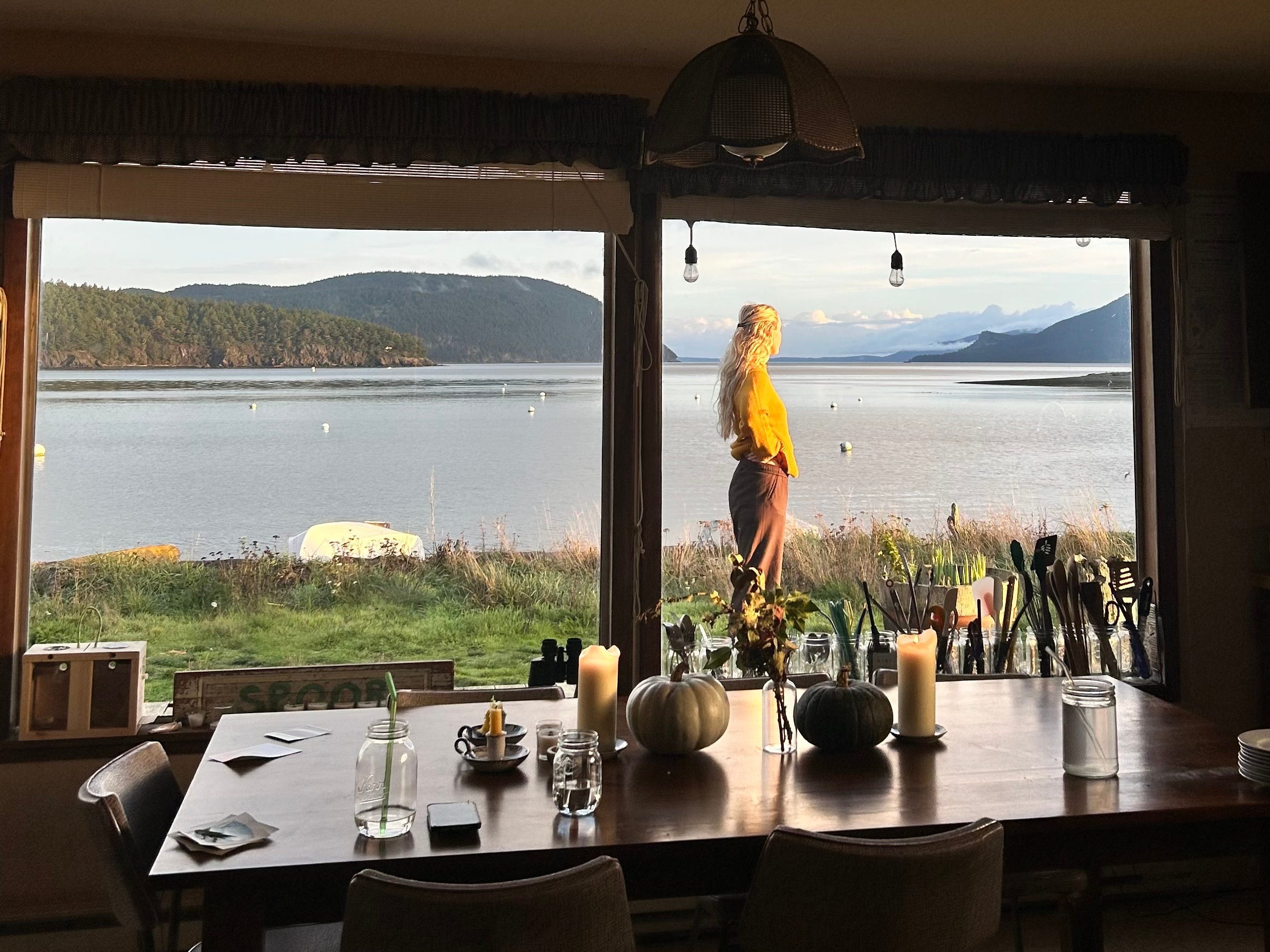A table near two large windows, with a view of Blakely standing outside near the water, with mountains in the background.