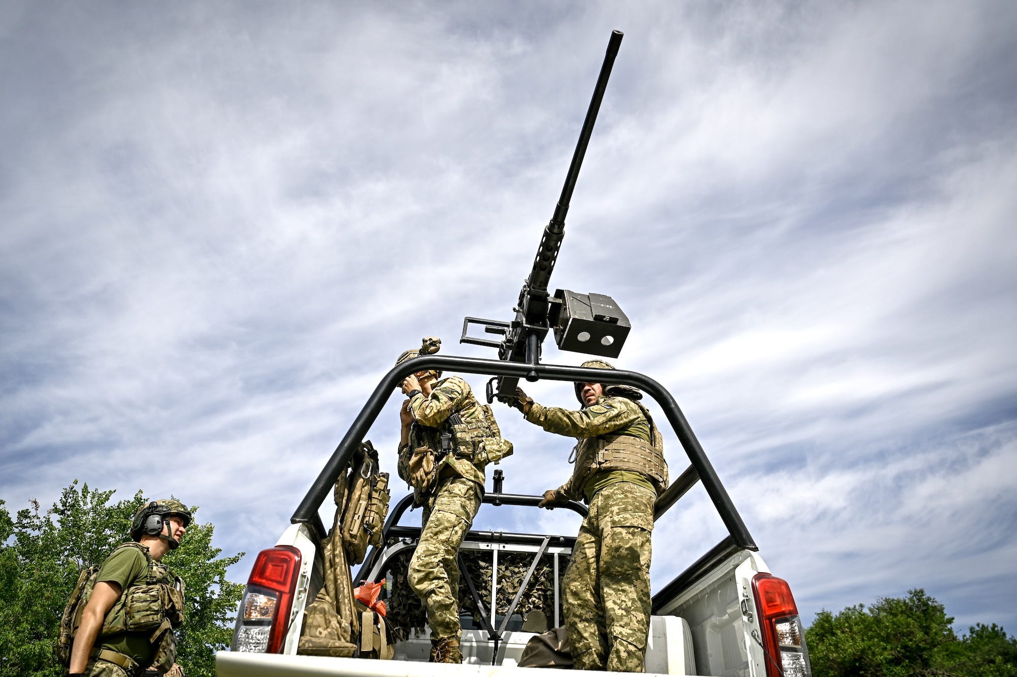 Two man in camoflage gear stand on the bed of a white truck with a large machine gun mounted on the back, under a cloudy blue sky
