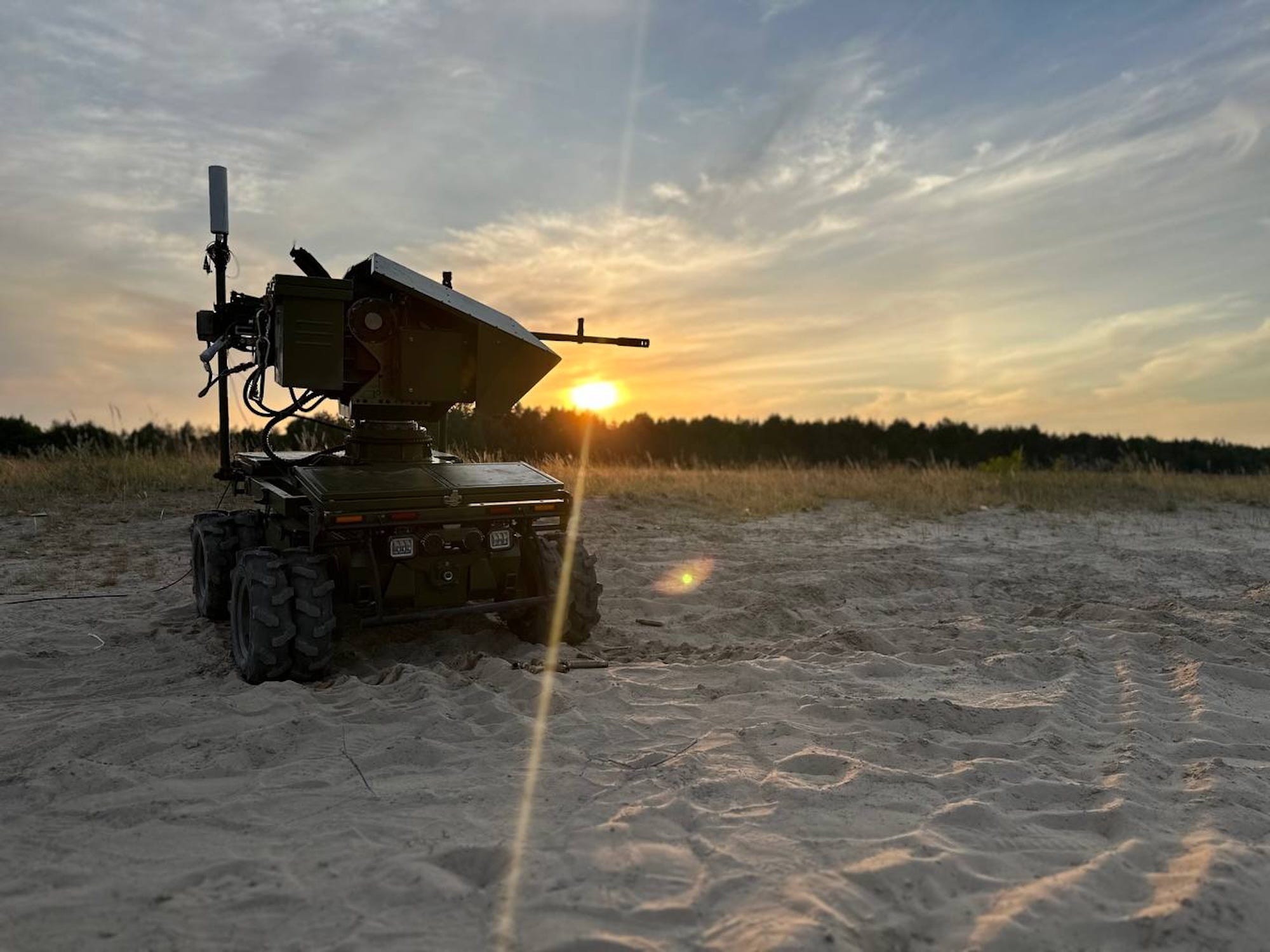 A large robot on tracks and with a machine gun on sand with a sunset or sunrise behind it