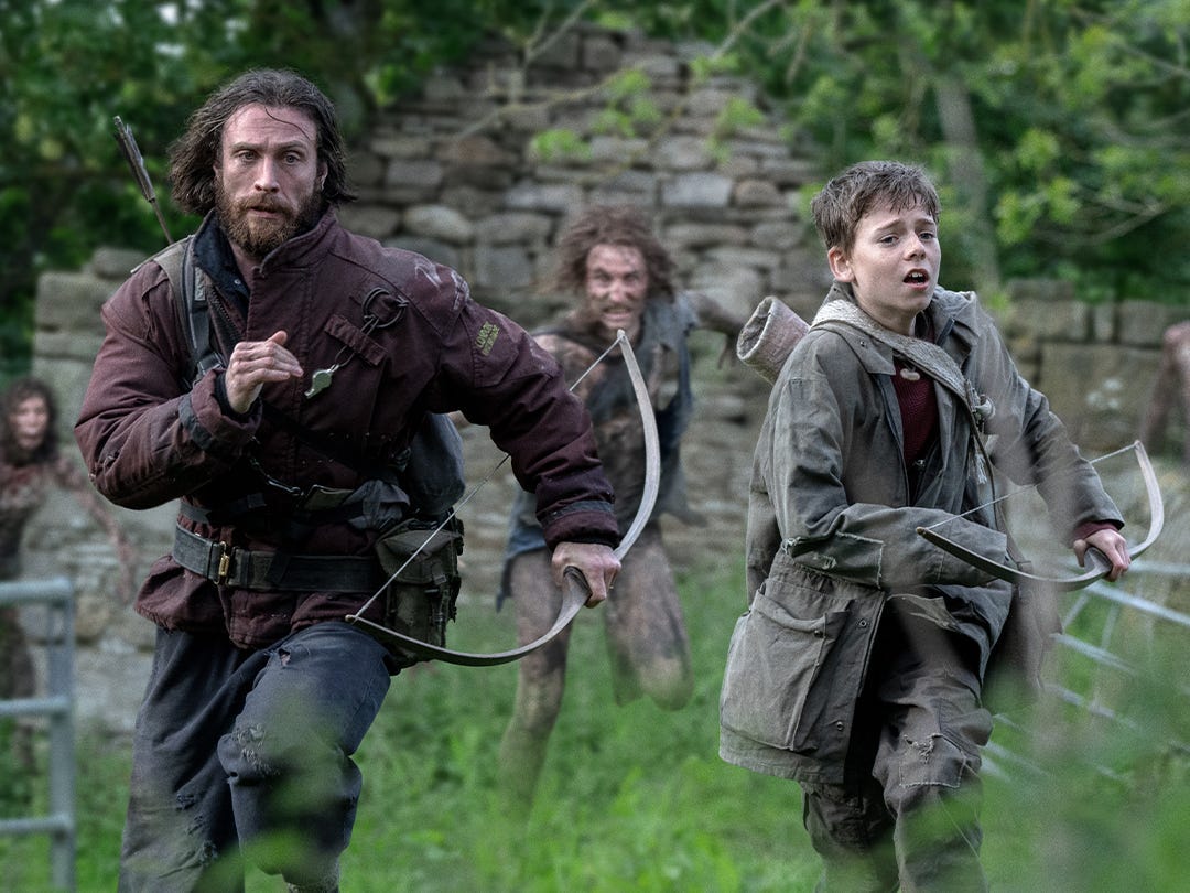 A man and a boy running away from other people behind them in a woodland area. Behind them is a wall made out of stones. On the left, the man has long brown hair and a long brown beard. He's wearing a burgundy coat with a quiver of arrows strapped to his back. He's holding a bow. On the right, the young boy wears a black green coat and has a white quiver on his back with strap across his chest. He's also holding a bow.