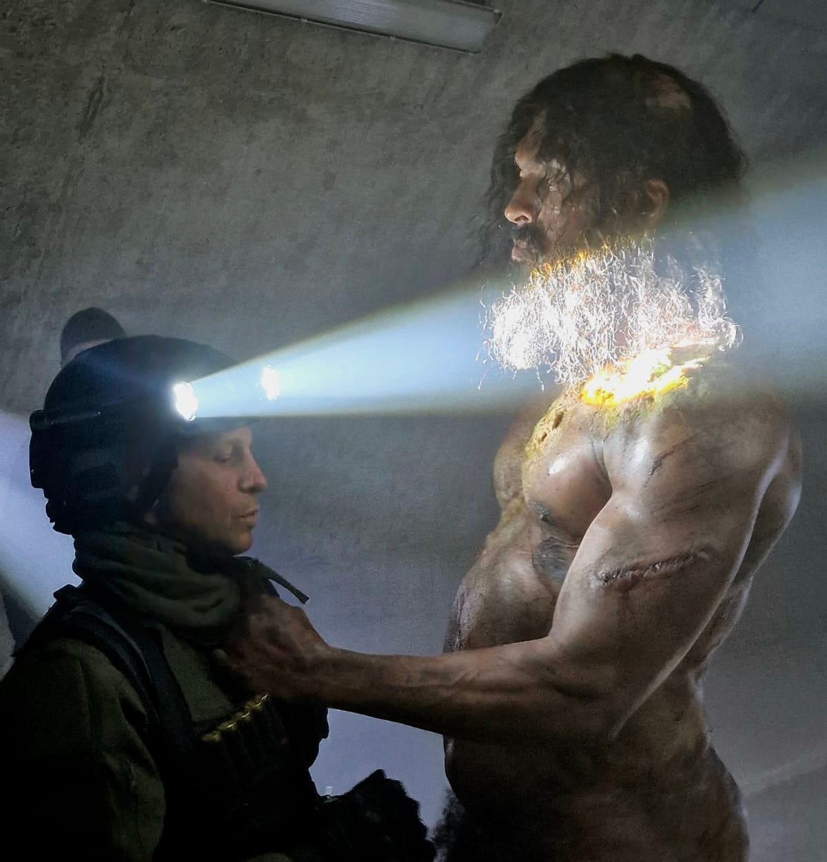 A naked man with long dark hair and a long beard is covered in deep scars and scratches. He's holding onto a soldier who is standing in front of him with his head torches illuminated.
