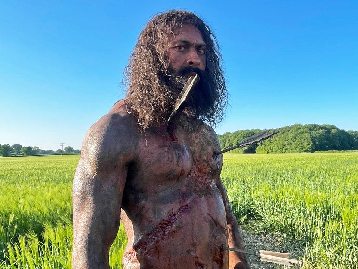 A photo of a naked man with long dark hair and a long beard wearing a prosthetic body that has three arrows sticking out of his chest and belly.