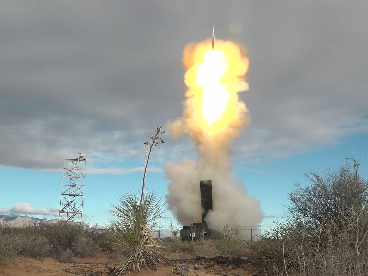A missile is fired from a launcher in a desert landscape with grey clouds across a blue sky in the background.