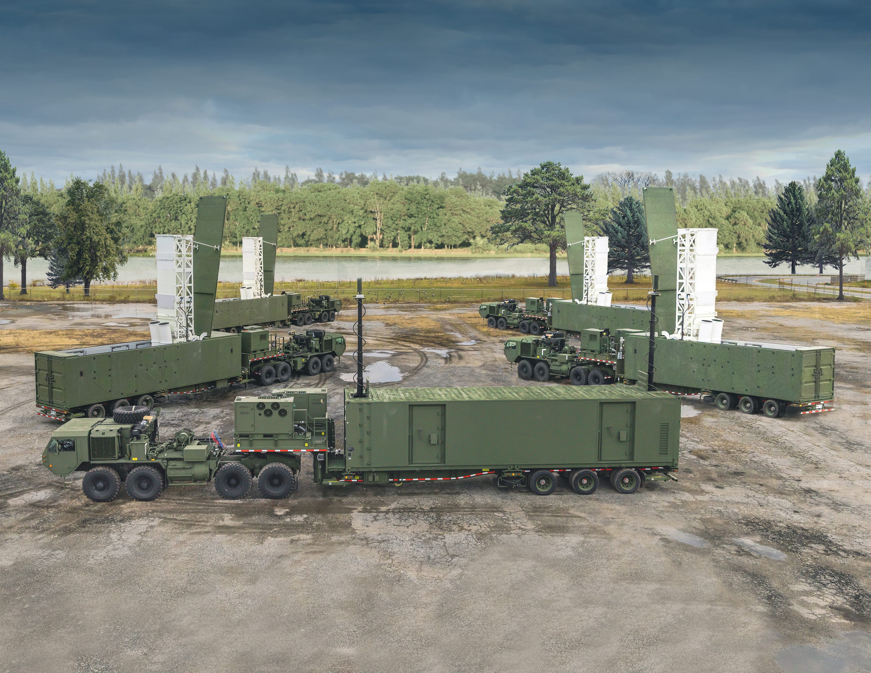 Four of the US Army's Mid-Range Capability missile systems, which are mounted on trucks, are parked in a formation on a dirt ground with trees and a grey sky in the background.