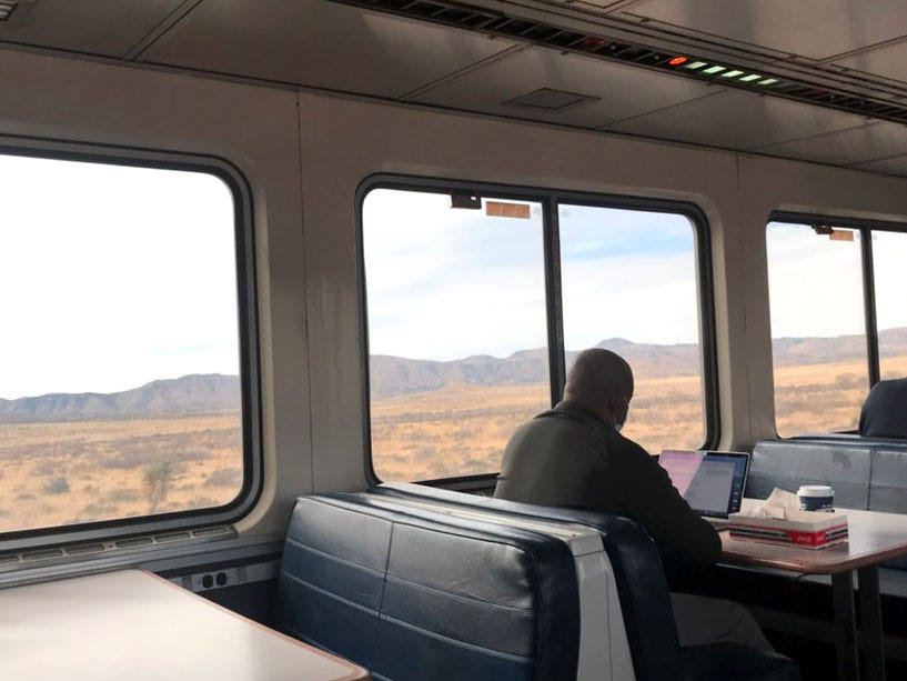 A man working on his laptop on a train, with a beautiful view of mountains and prairies out the window.