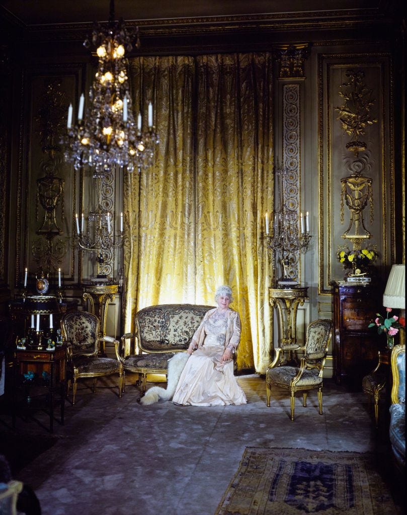 Mrs. Cornelius Vanderbilt seated in the classic Louis XVI-styled parlor of the Fifth Avenue Vanderbilt mansion, smiling for the camera.