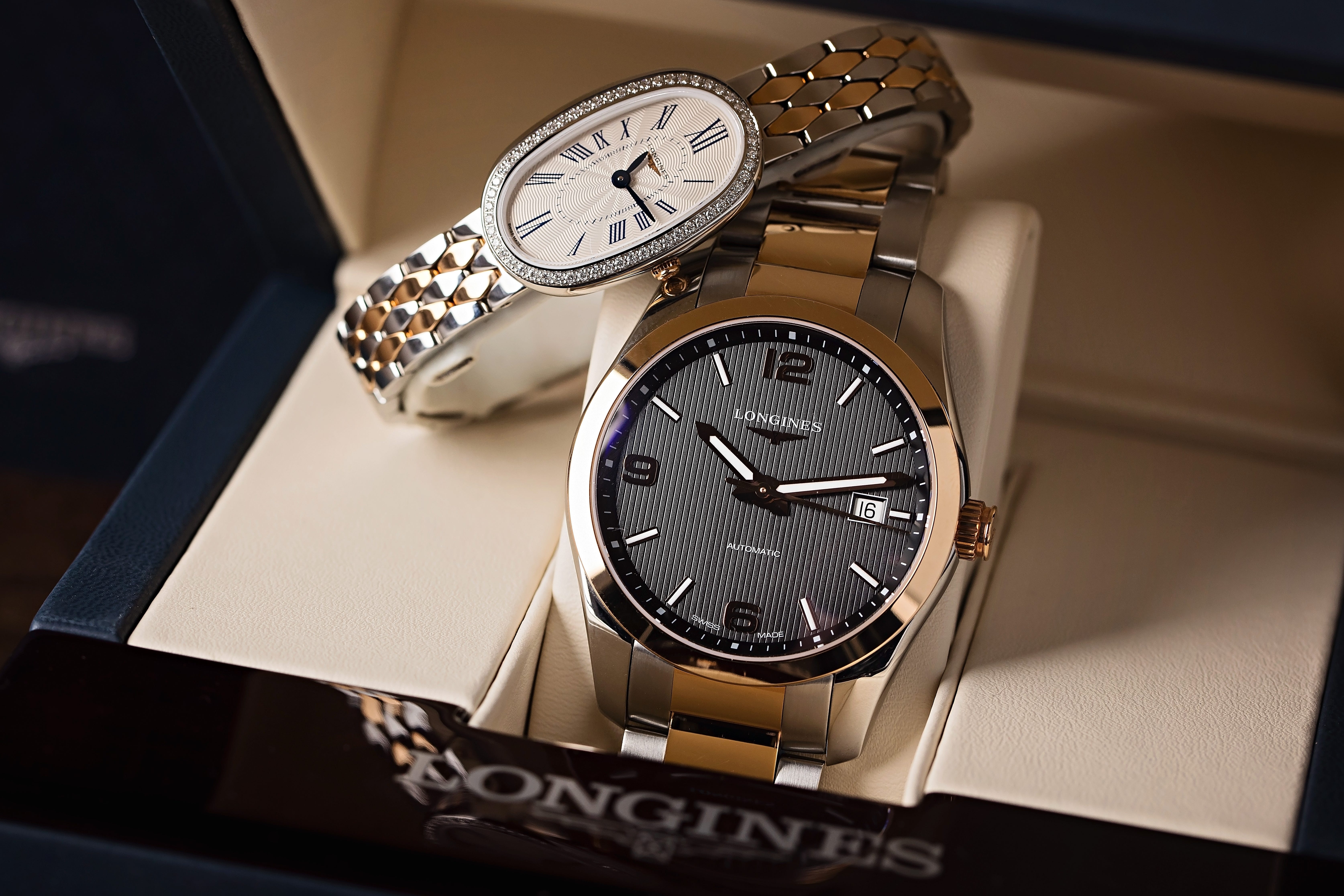 Two Longines watches in a box, one with a thin strap and an oval fave and one with a thicker strap and round face.