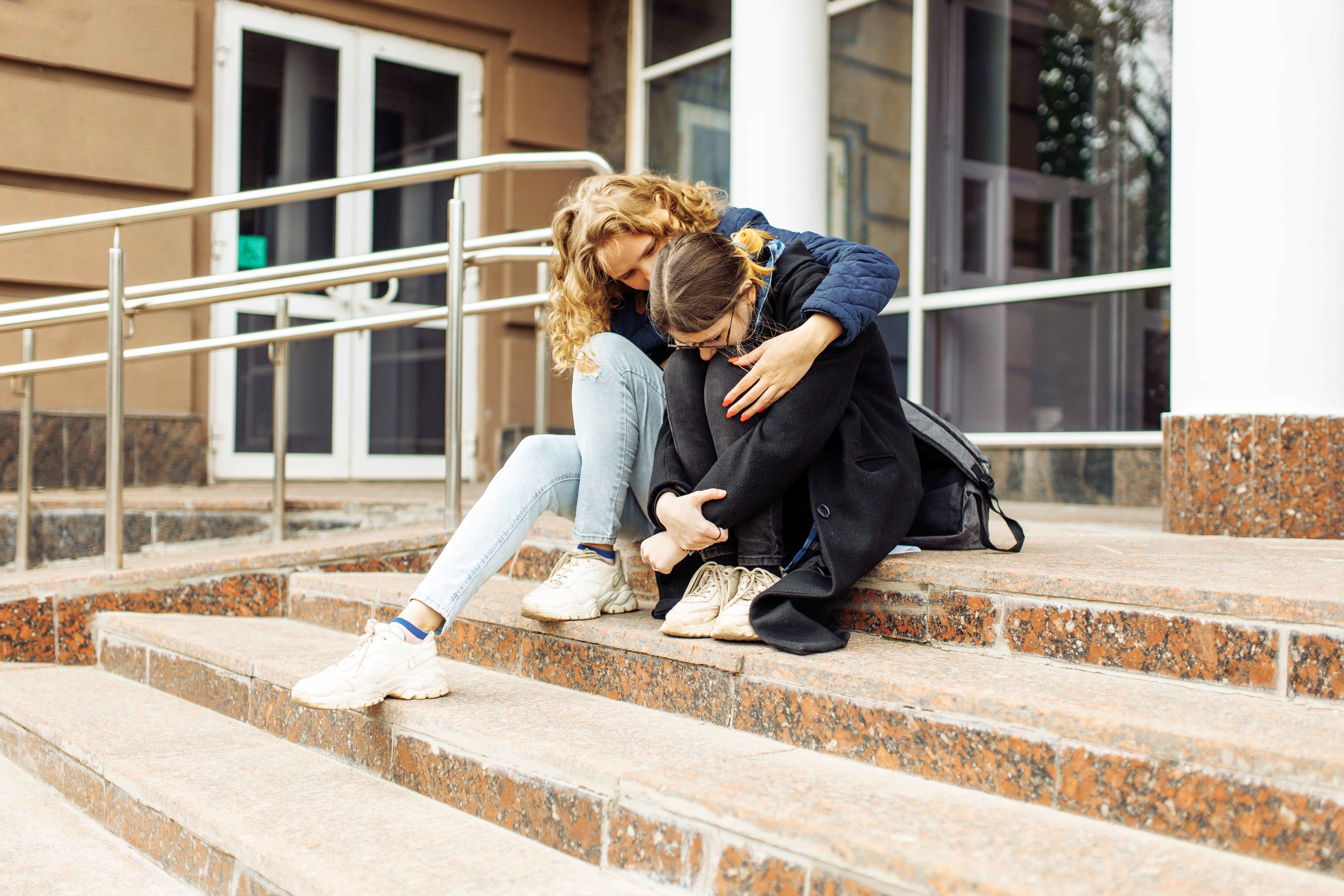 A woman comforts a girl while sitting on steps outside of a building.