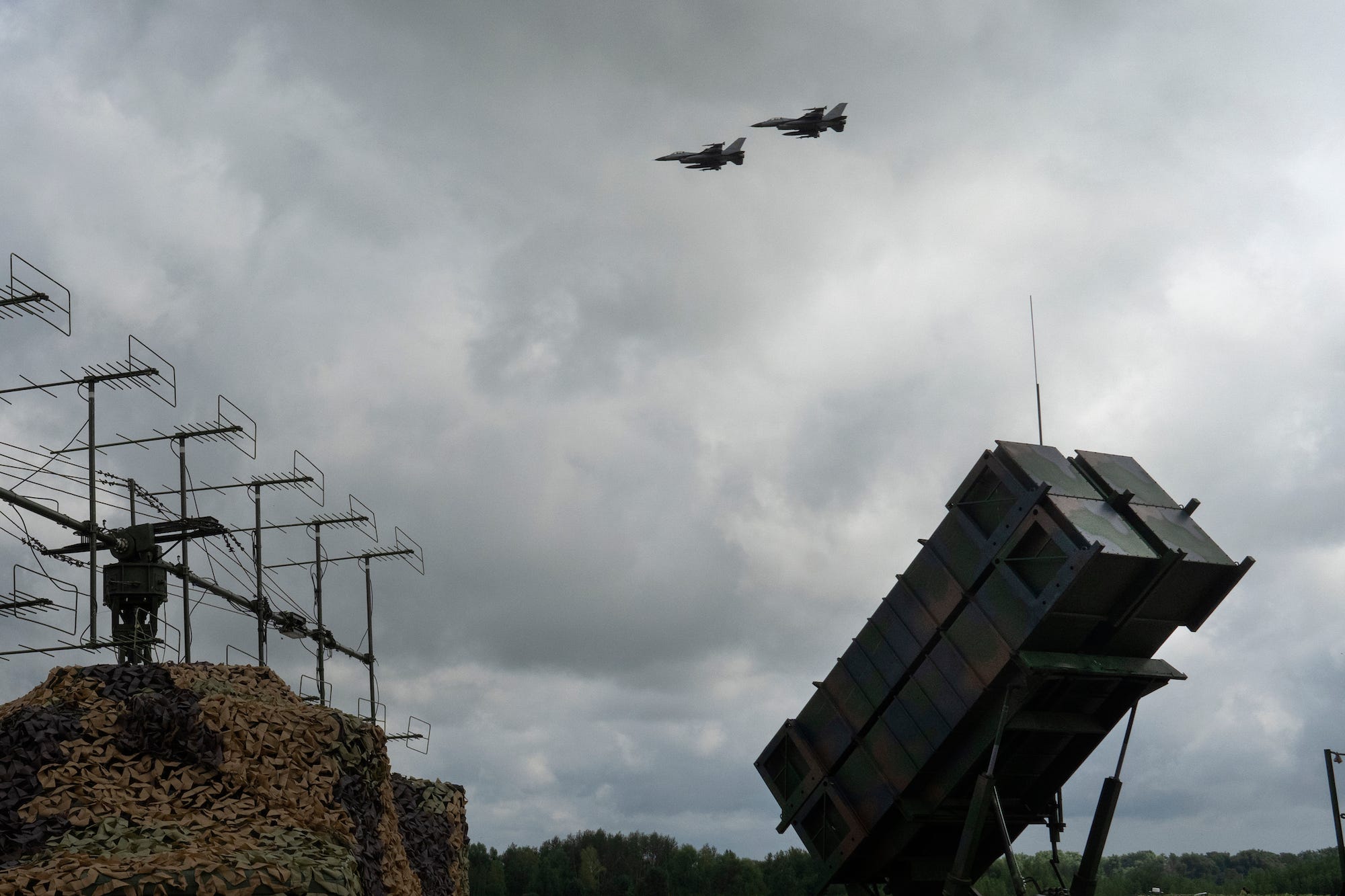 Two F-16 fighter jets fly over a Patriot Air and Missile Defense System against a gray sky