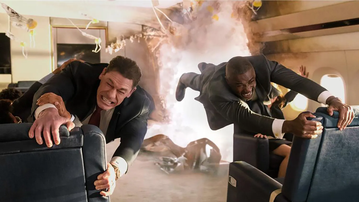 John Cena and Idris Elba hold on to seats on a plane, a hole in the plane is seen behind them.