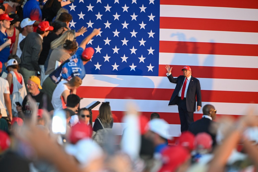 Donald Trump waving to a crowd at a rally in front of a large American flag.