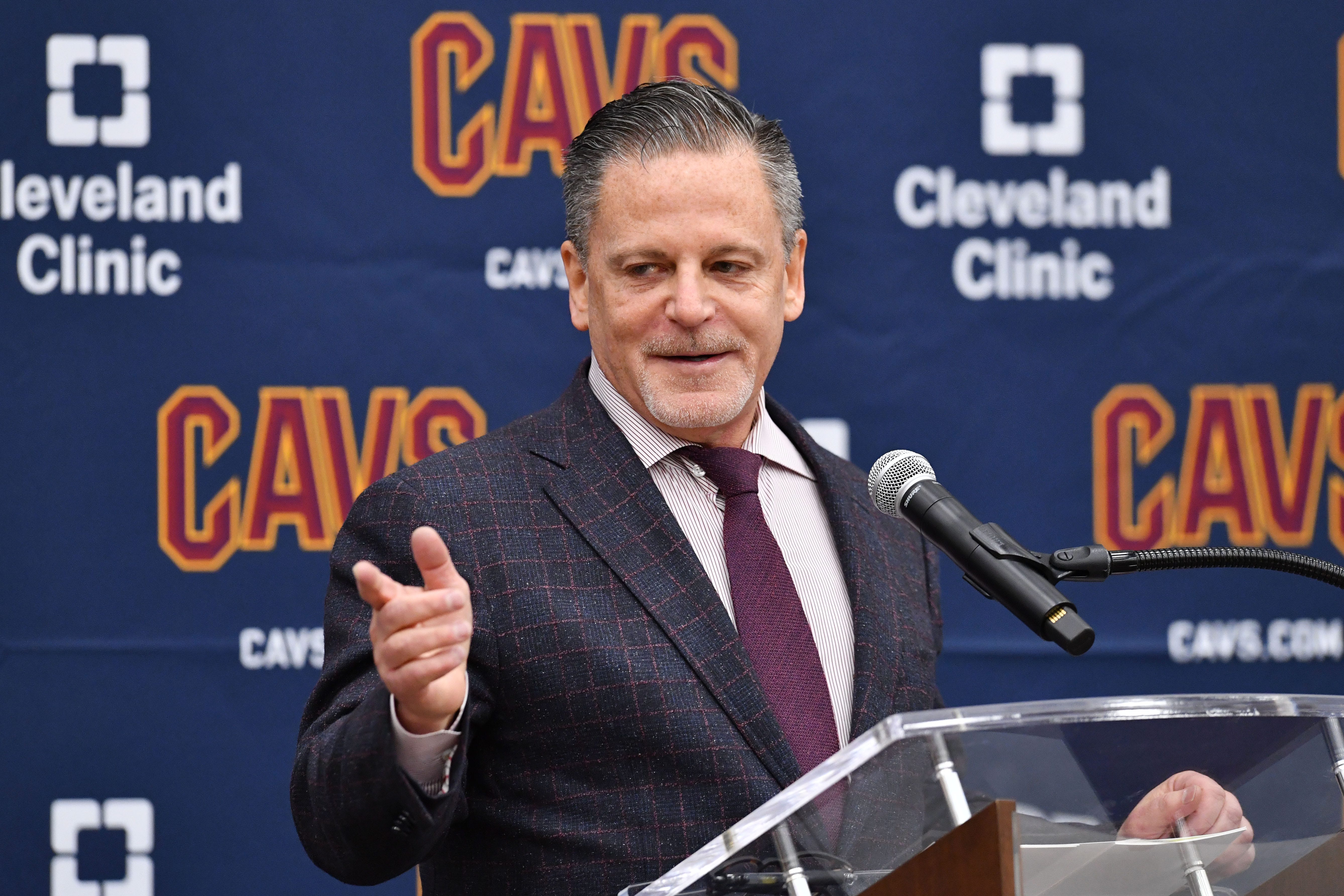 Dan Gilbert behind a microphone with a blue Cleveland Cavaliers backdrop with Cleveland Clinic logos on it