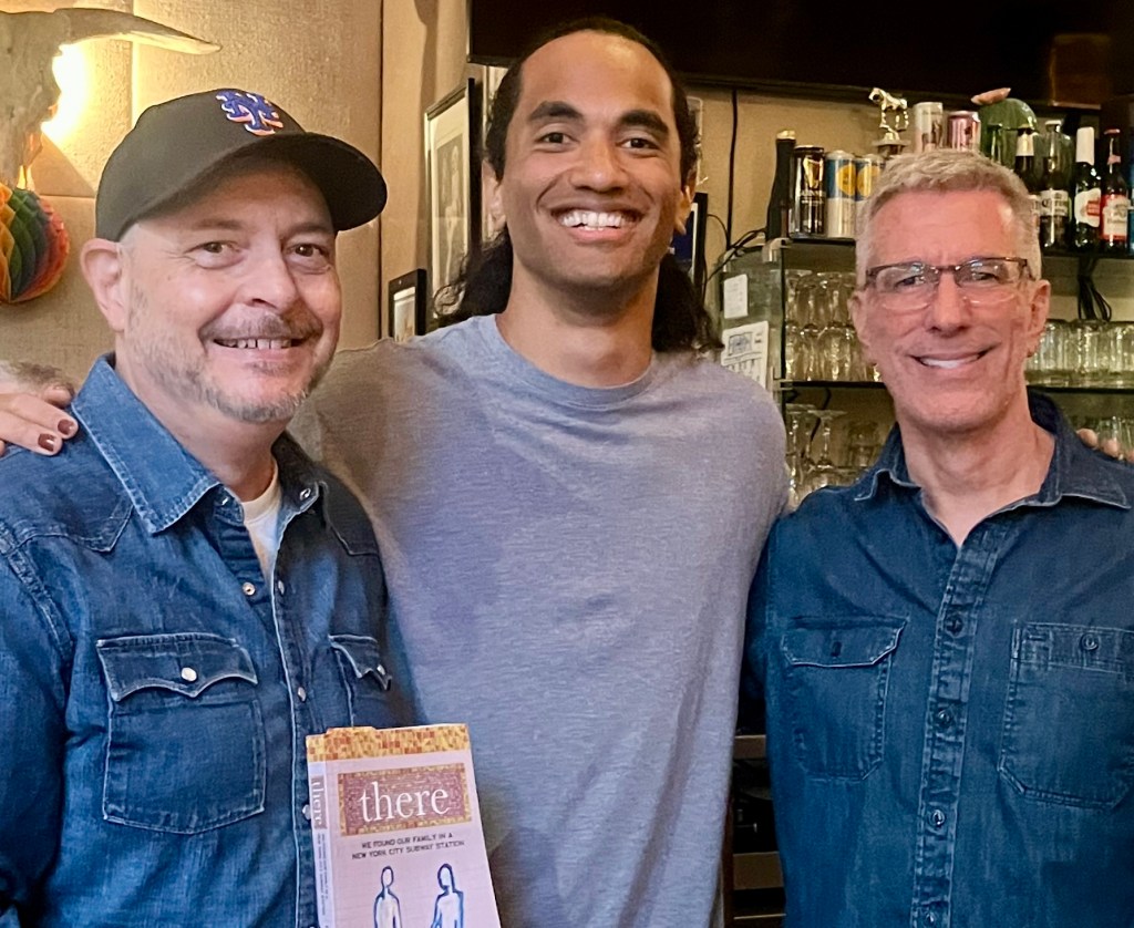 Peter Mercurio, holding a book, his adult son Kevin and husband Danny Stewart stand in front of a bar.