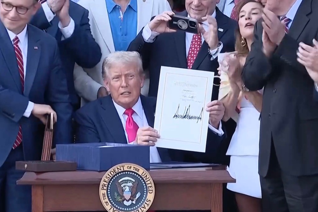 President Trump signing a document at the White House, surrounded by onlookers.