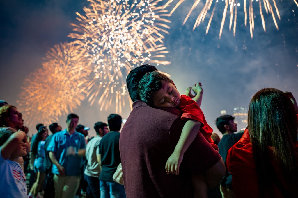 A child sleeping on his father's shoulder during a fireworks display.