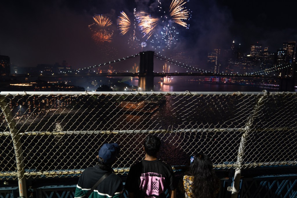 Fireworks over the Brooklyn Bridge, viewed from behind a chain-link fence by several people.