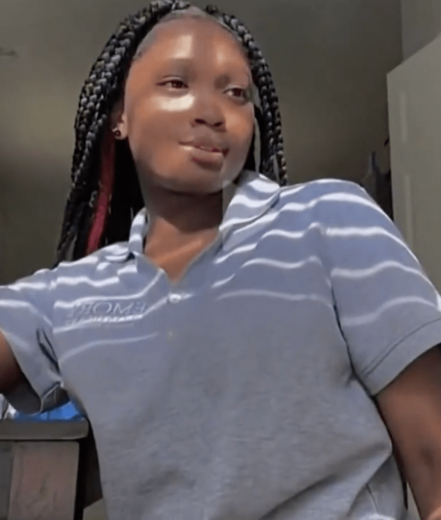 Screenshot of a young woman with long braids wearing a striped polo shirt.