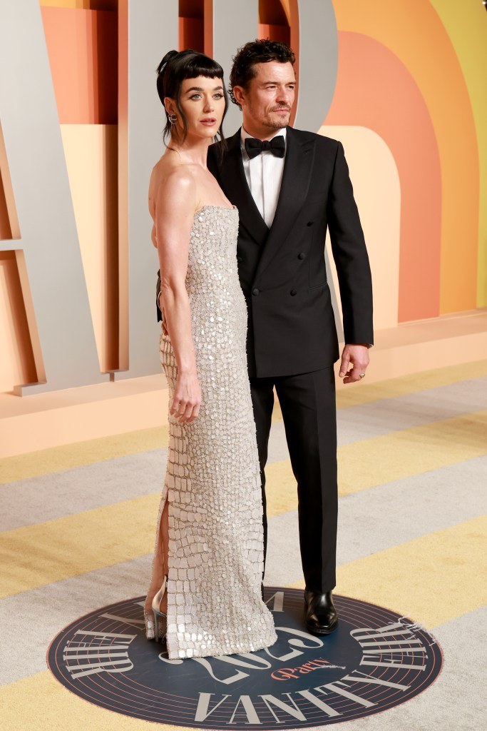 Katy Perry in a white sequins strapless dress holding hands with Orlando Bloom who is in a bow tie and suit on a red carpet. 