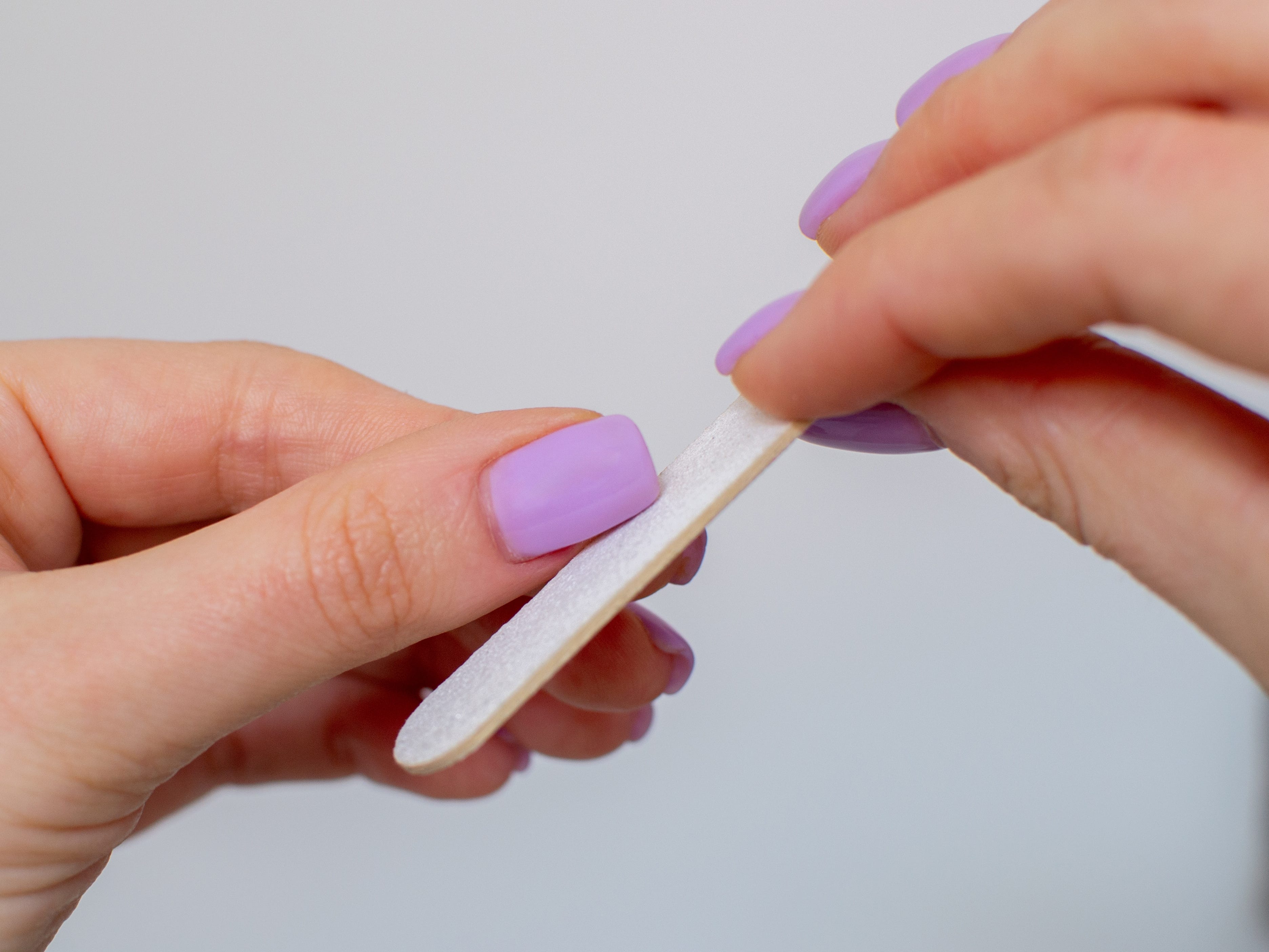 Close-up of purple nails, as the right hand holding a nail file shapes the thumbnail of the left hand.