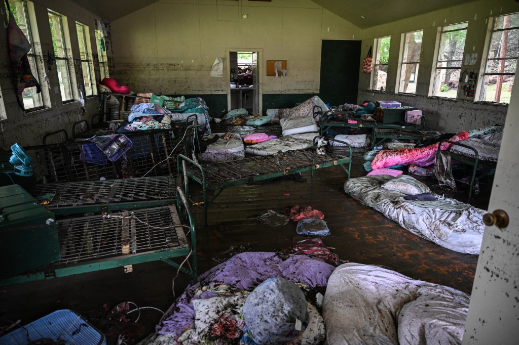 Muddy interior of a flood-damaged cabin with overturned beds and belongings.